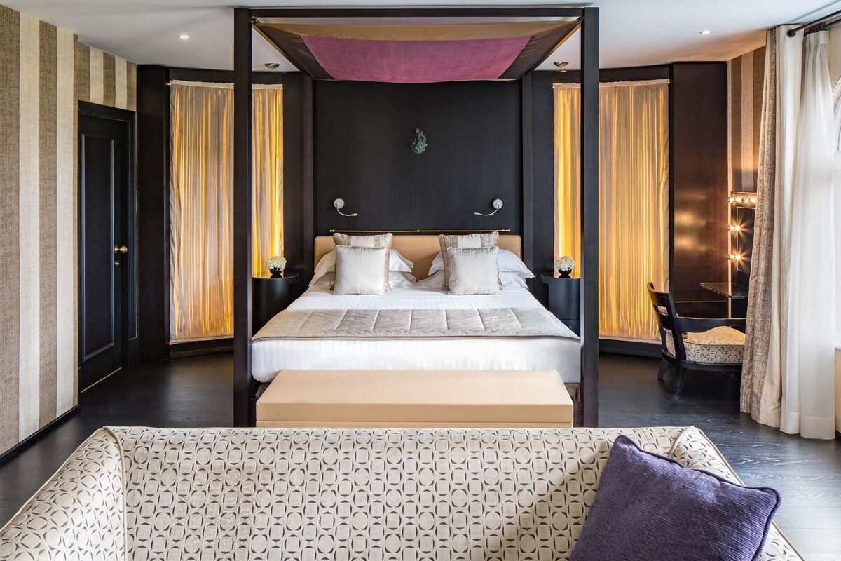 A modern hotel room with a large bed, a sitting area with a patterned sofa, and a vanity with a mirror and lights.