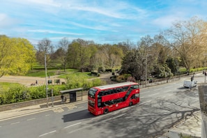 A red double-decker bus on a city street with a park and trees in the background. - Delano London (London)