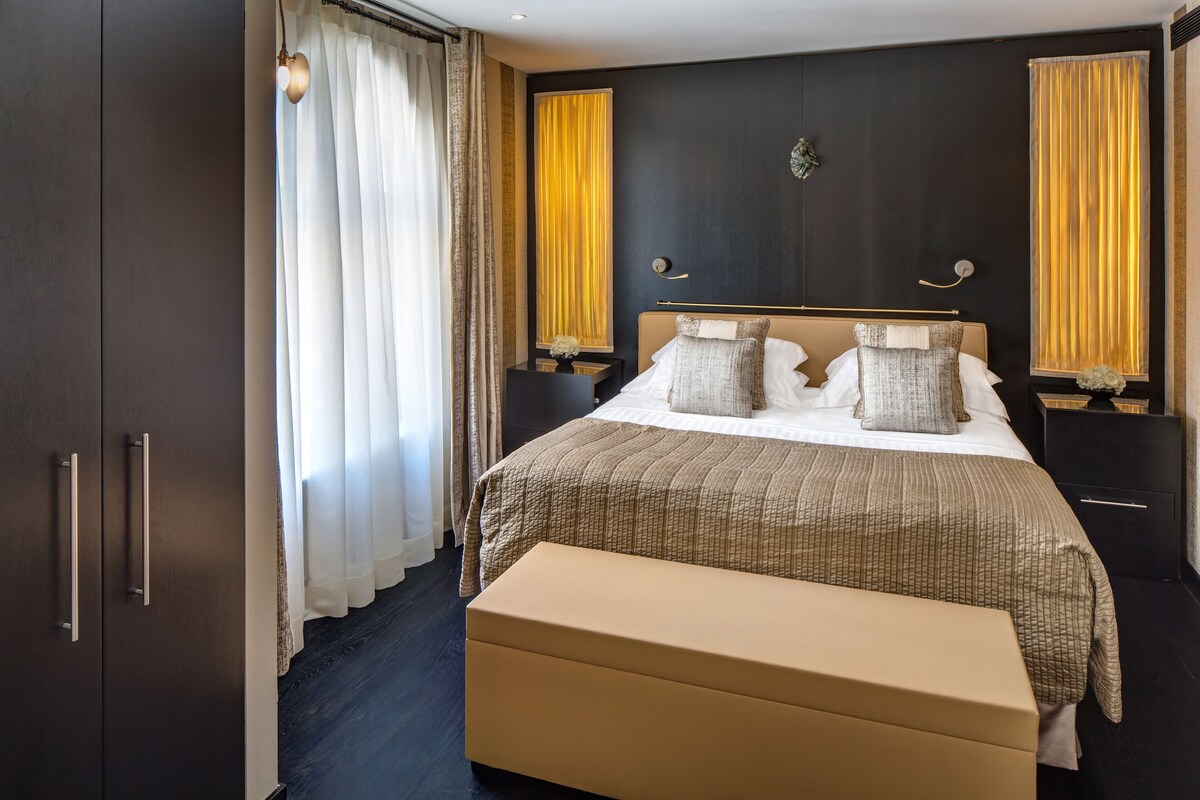 A modern hotel room with a large bed, a bench, and a dark wooden wardrobe.