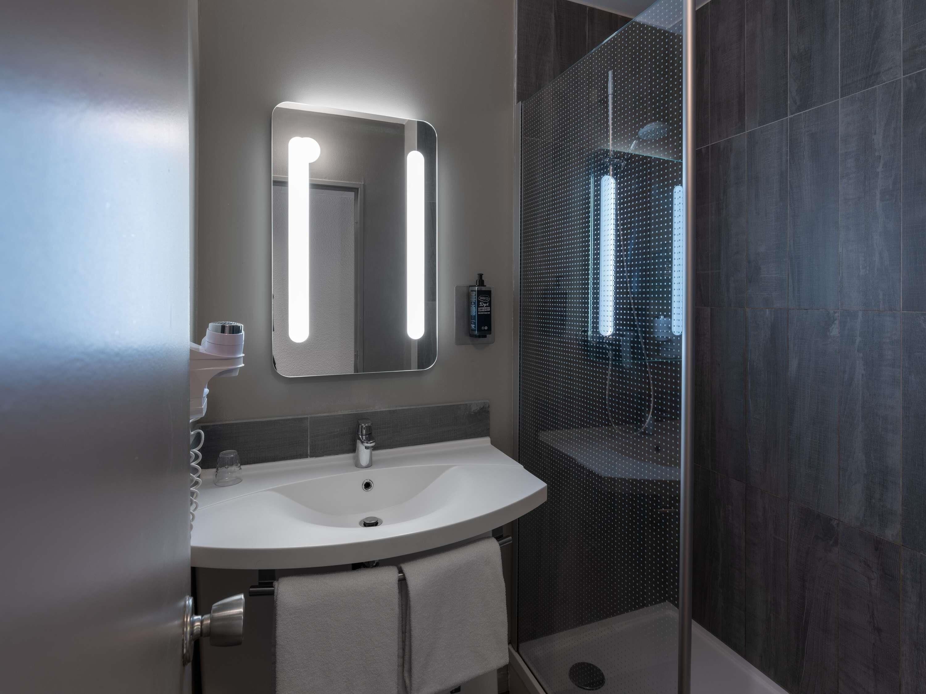 A modern bathroom with a white sink, a mirror with backlighting, a handheld hair dryer, and a glass shower enclosure.