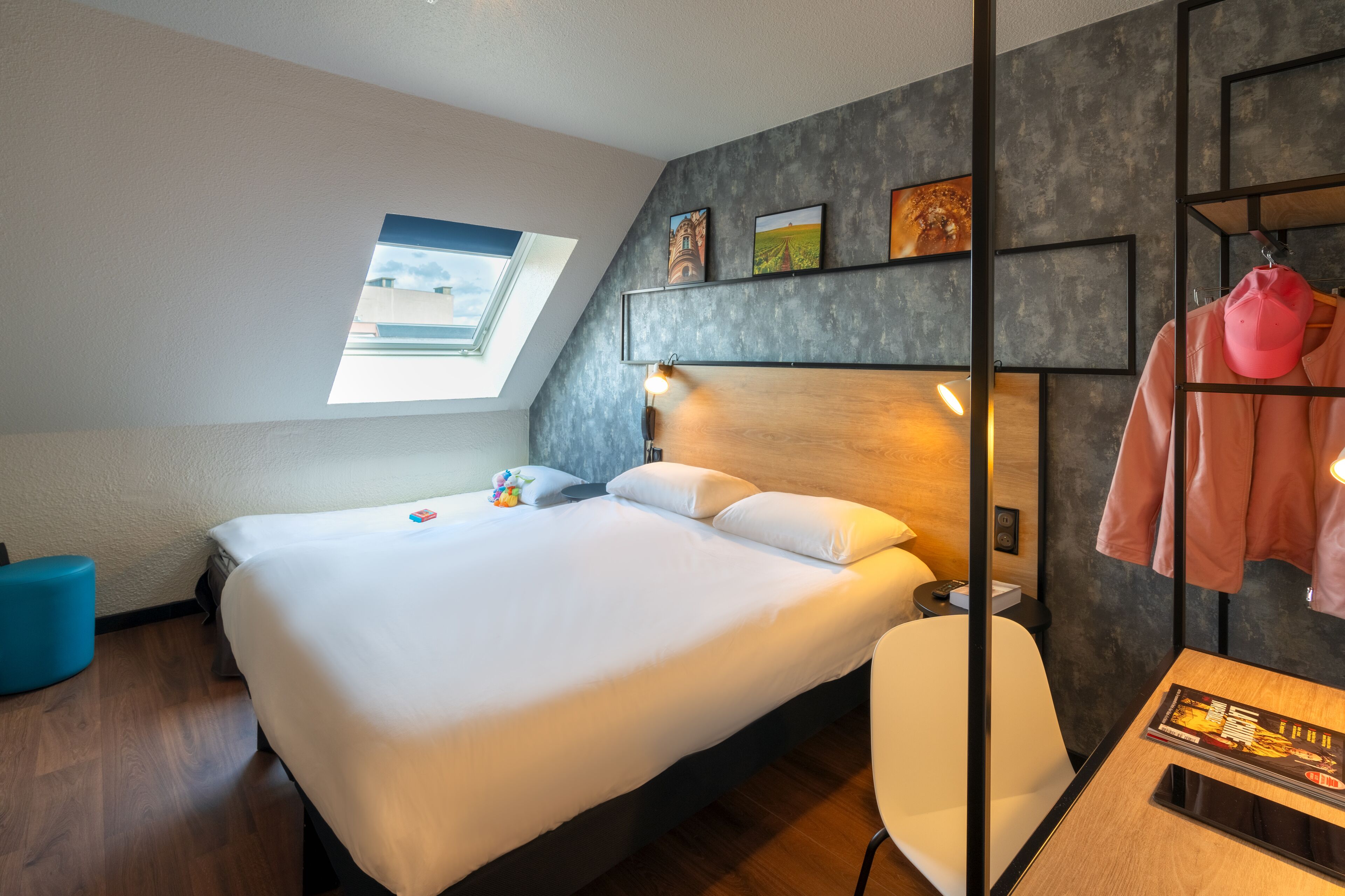 Una habitación de hotel moderna con una cama grande, un tragaluz, un cabecero de madera, una mesita de noche con una revista y un otoman azul.