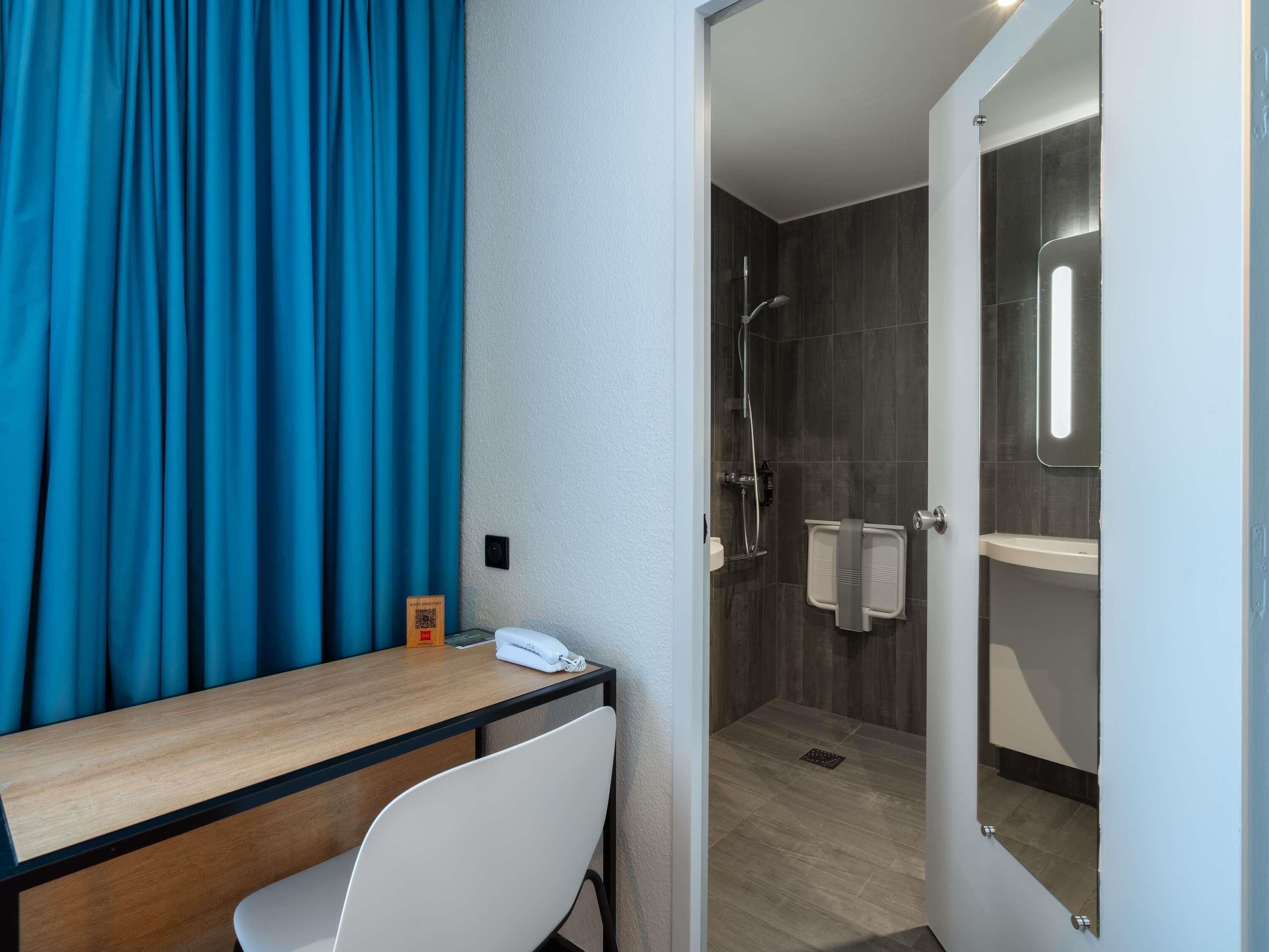 Une chambre d’hôtel avec un bureau en bois, une chaise blanche, un espace douche doté d’une porte en verre et un rideau bleu.