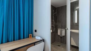 Une chambre d’hôtel avec un bureau en bois, une chaise blanche, un espace douche doté d’une porte en verre et un rideau bleu.