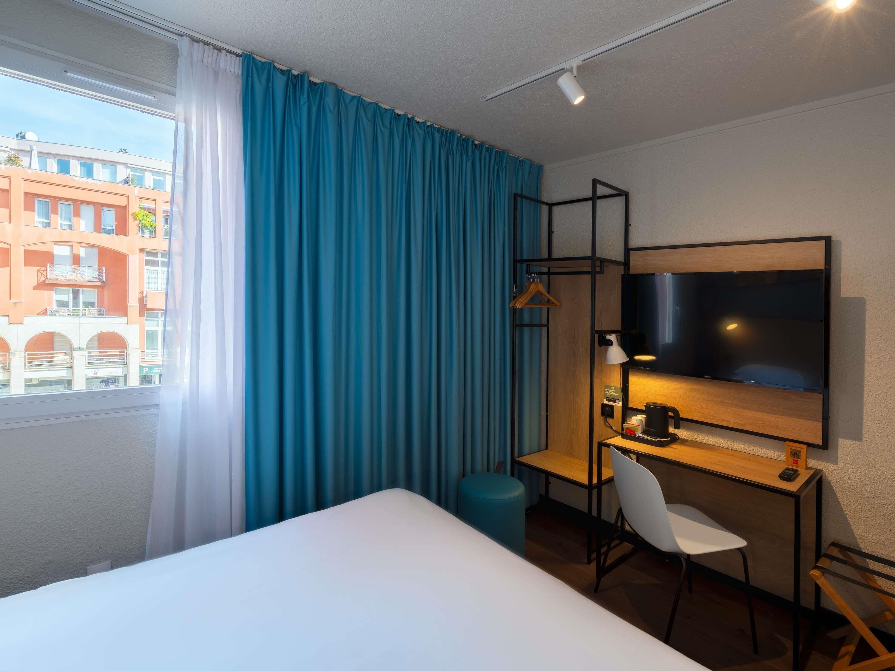 A hotel room with a bed, a desk with a chair, a TV, and a window with curtains.