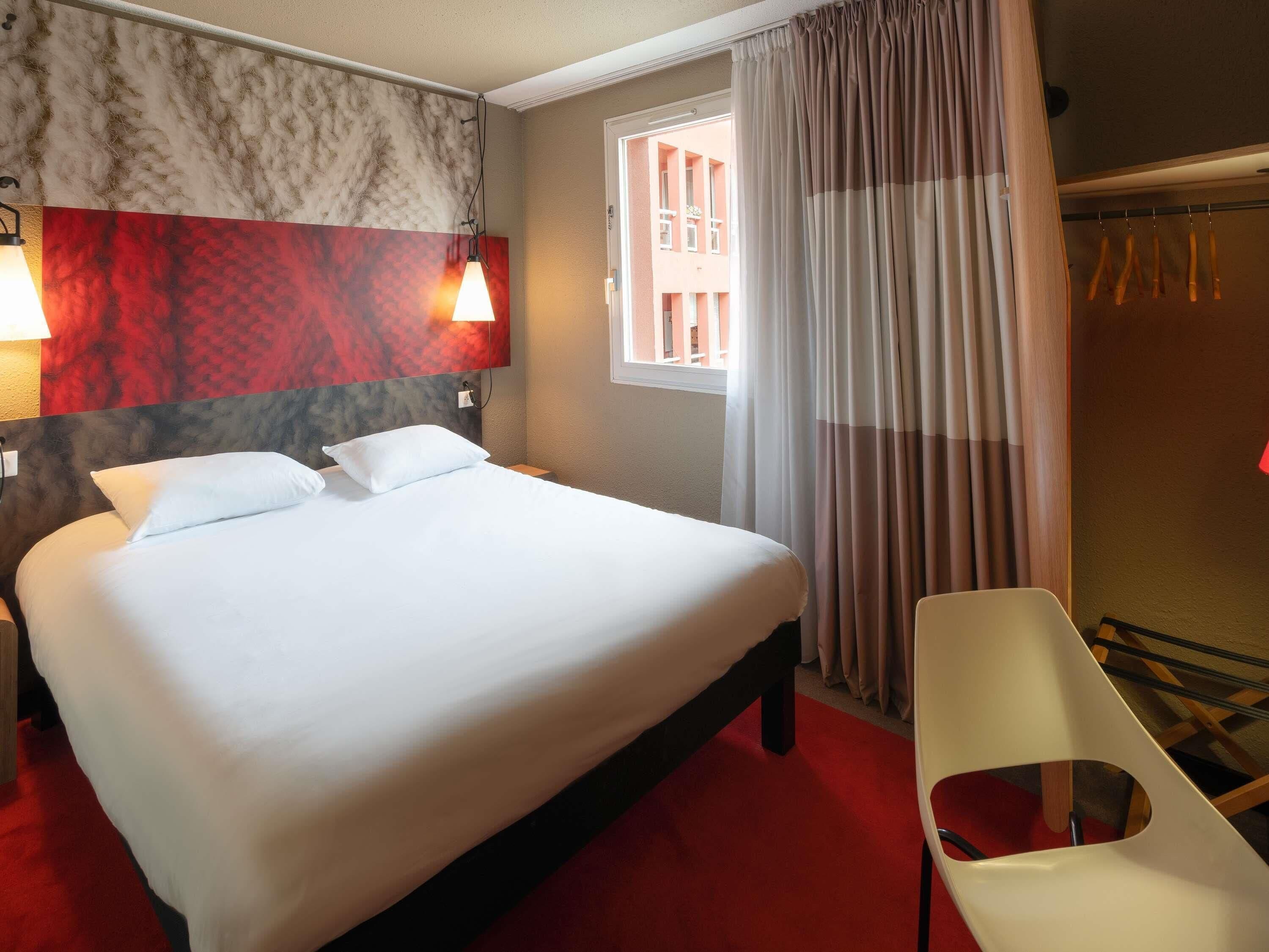Una camera d'albergo con un letto, due abat-jour, una finestra con tende, una sedia e uno spazio doccia.
