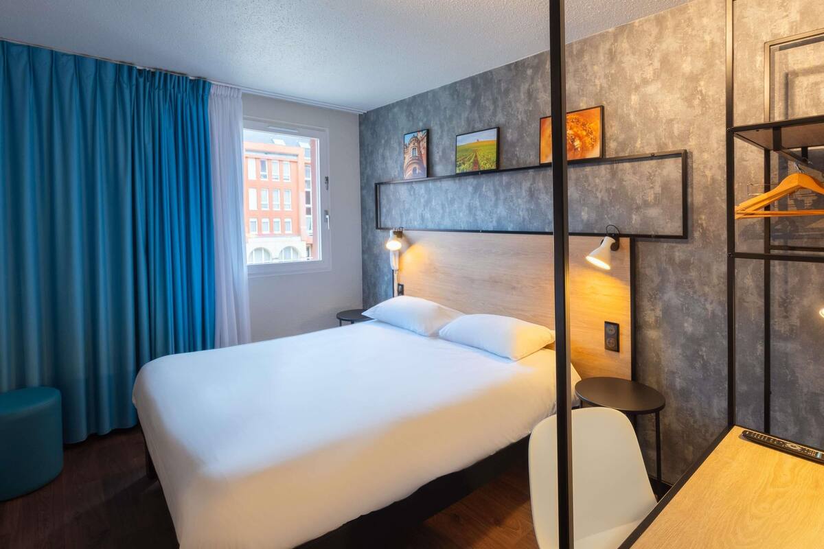 Ein modernes Hotelzimmer mit einem großen Bett, einem Schreibtisch und einem Stuhl. Es gibt ein Fenster mit Vorhängen, eine Wandablage mit Bildern und eine Tür mit Kleiderhaken.