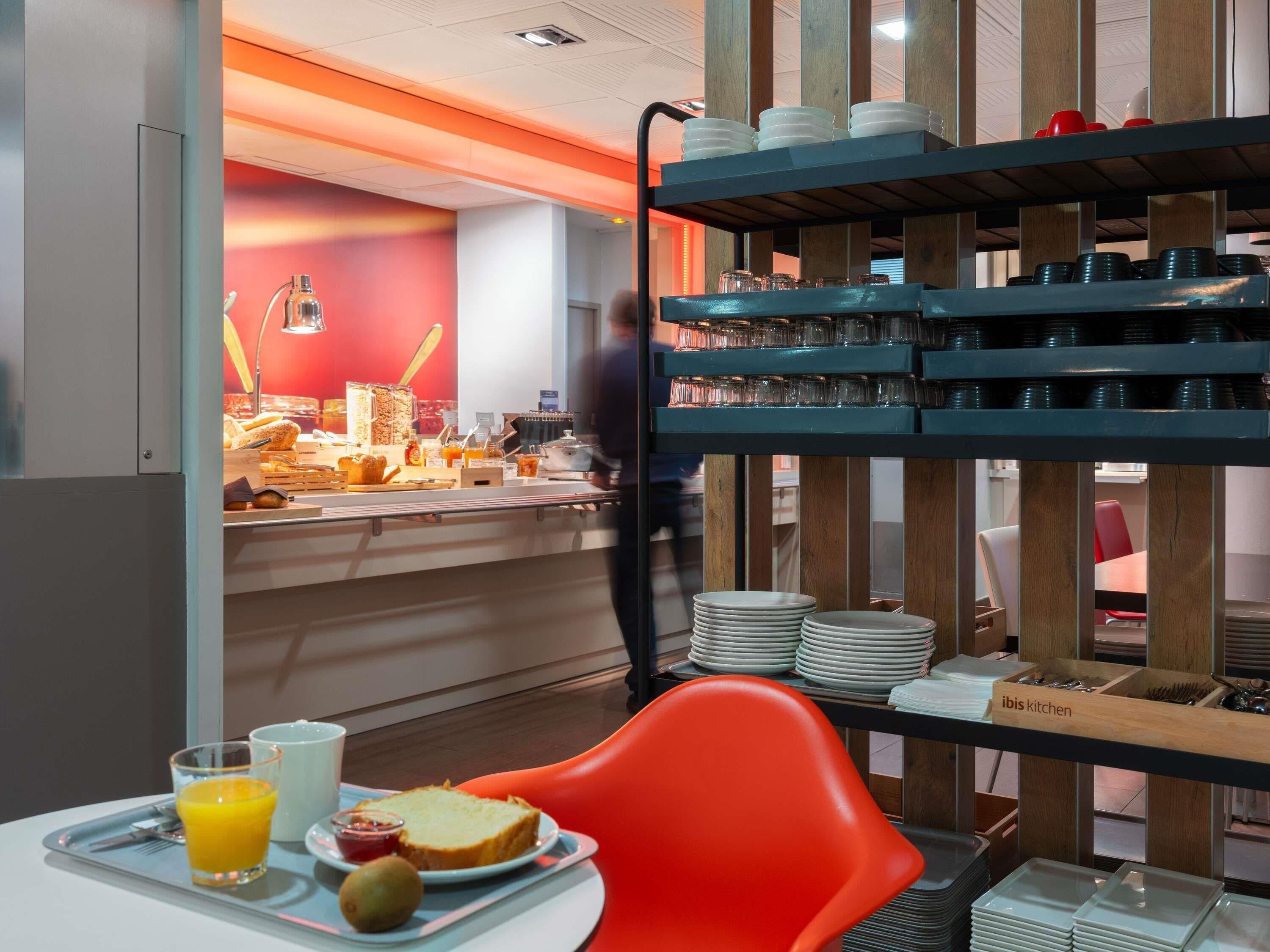 Een ontbijtbuffet met diverse soorten eten, een persoon op de achtergrond en een feloranje stoel.