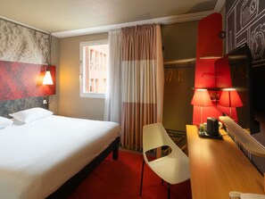 Quarto de hotel com cama, escrivaninha com luminária, cadeira e vista para o exterior.