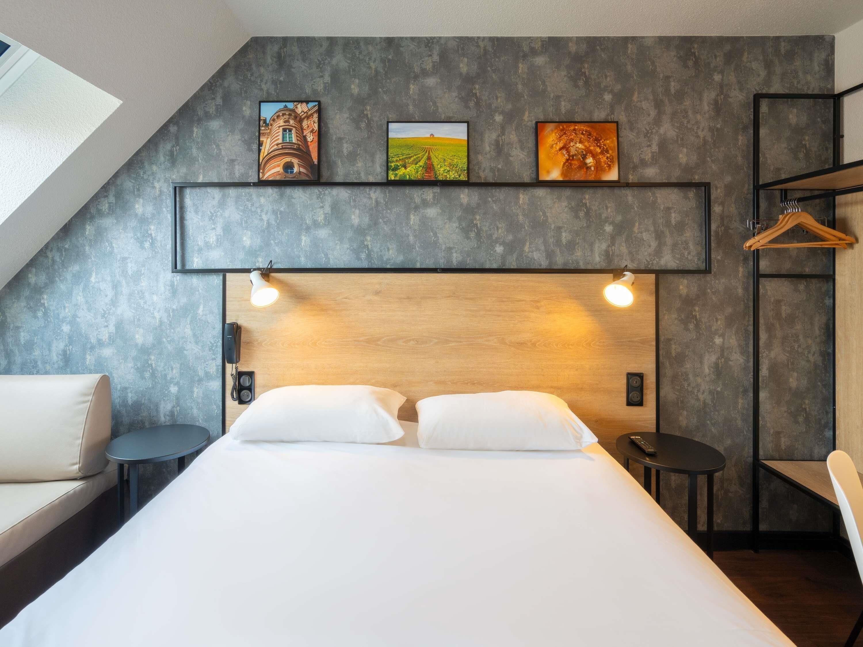 Habitación de hotel moderna con techo inclinado, una cama con dos almohadas blancas, una mesita redonda pequeña, una silla y un cabecero de madera.