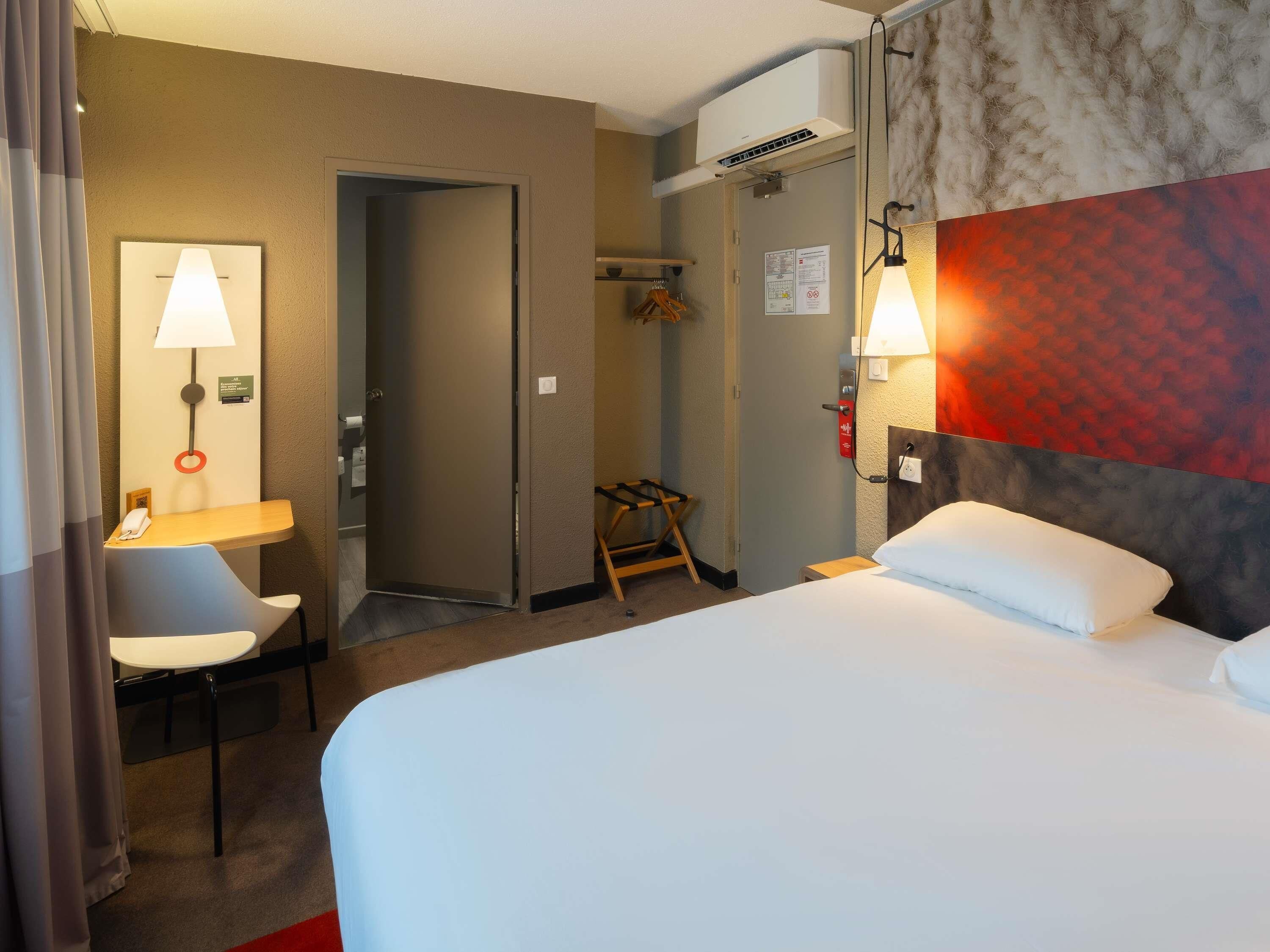 Een hotelkamer met een bed, een bureau met stoel, een lamp, een airconditioning en een deur.