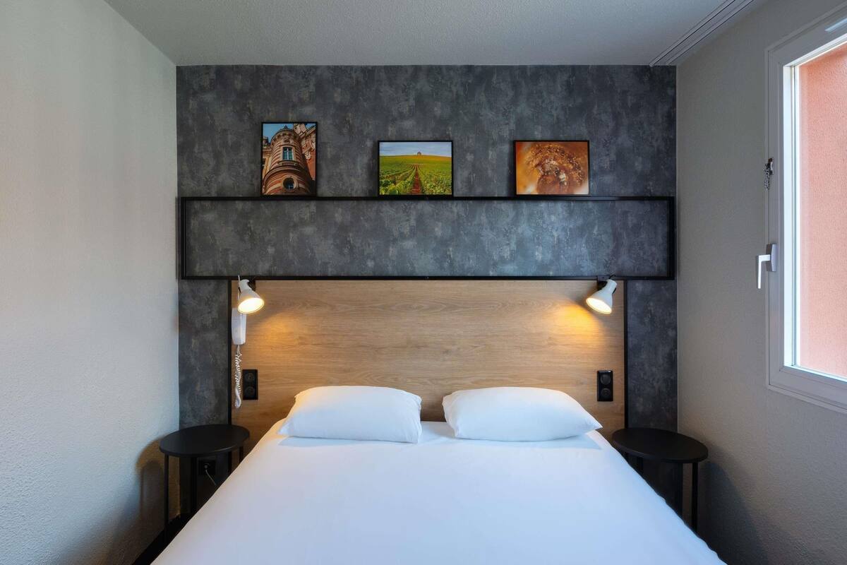 Ein modernes Hotelzimmer mit Bett, Nachttischen, einem Kopfteil mit integrierter Beleuchtung und einem Bild an der Wand.