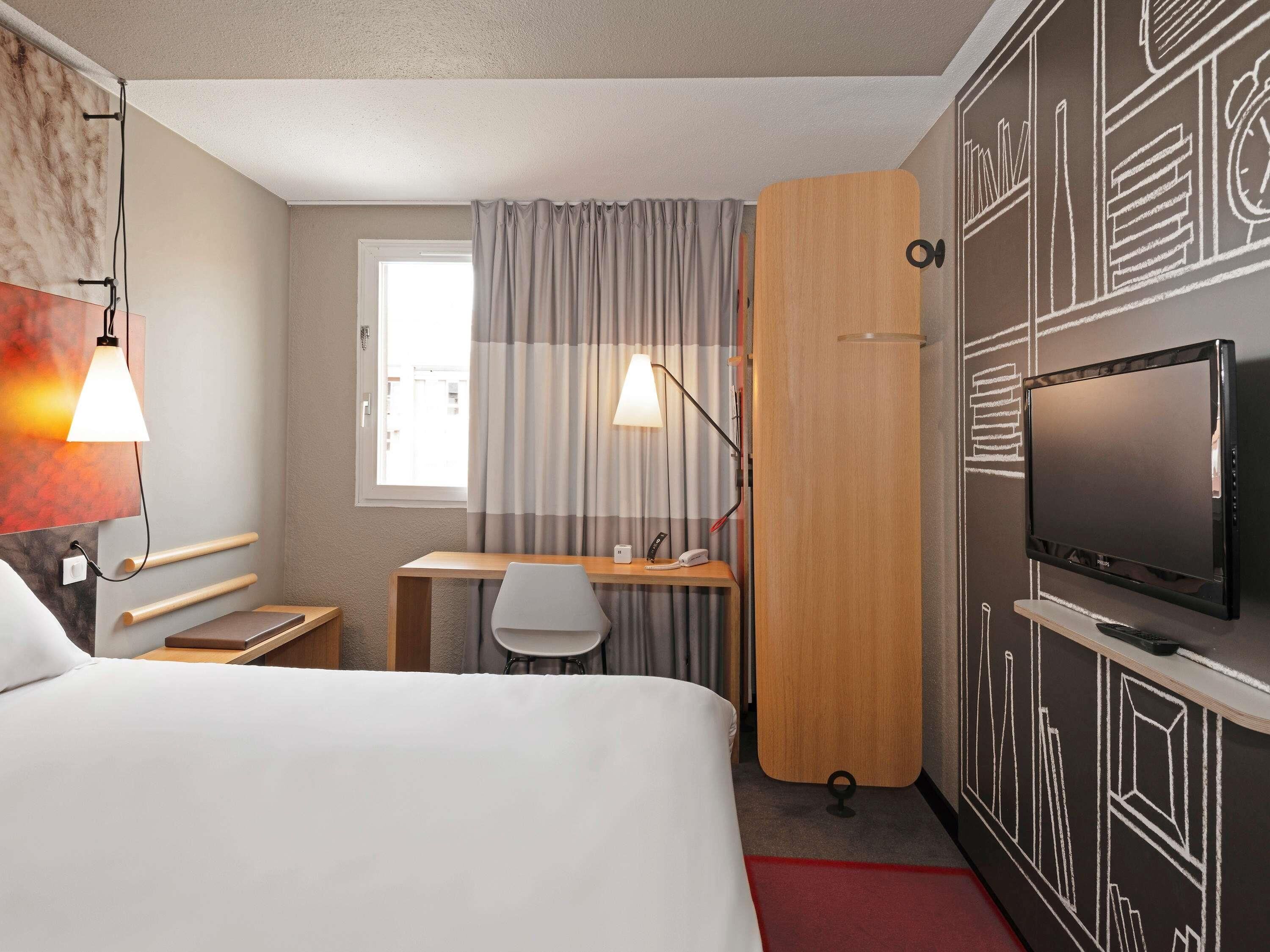 Habitació d'hotel amb llit, escriptori, cadira, televisió i finestra amb cortines.