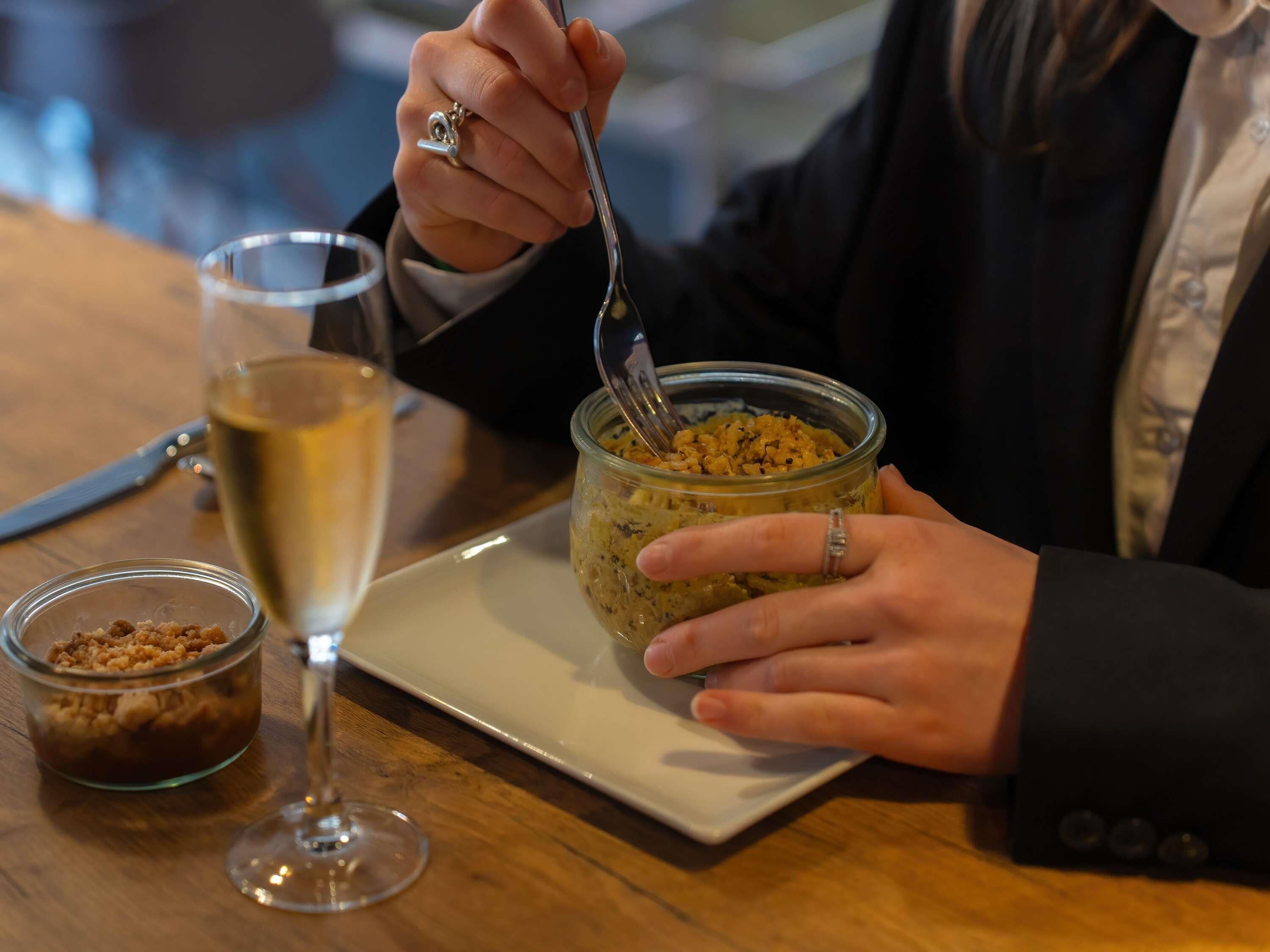 Una persona comiendo un postre de un tarro de vidrio con un tenedor, una copa de vino blanco y un cuenco pequeño con cobertura desmenuzada sobre una mesa de madera.