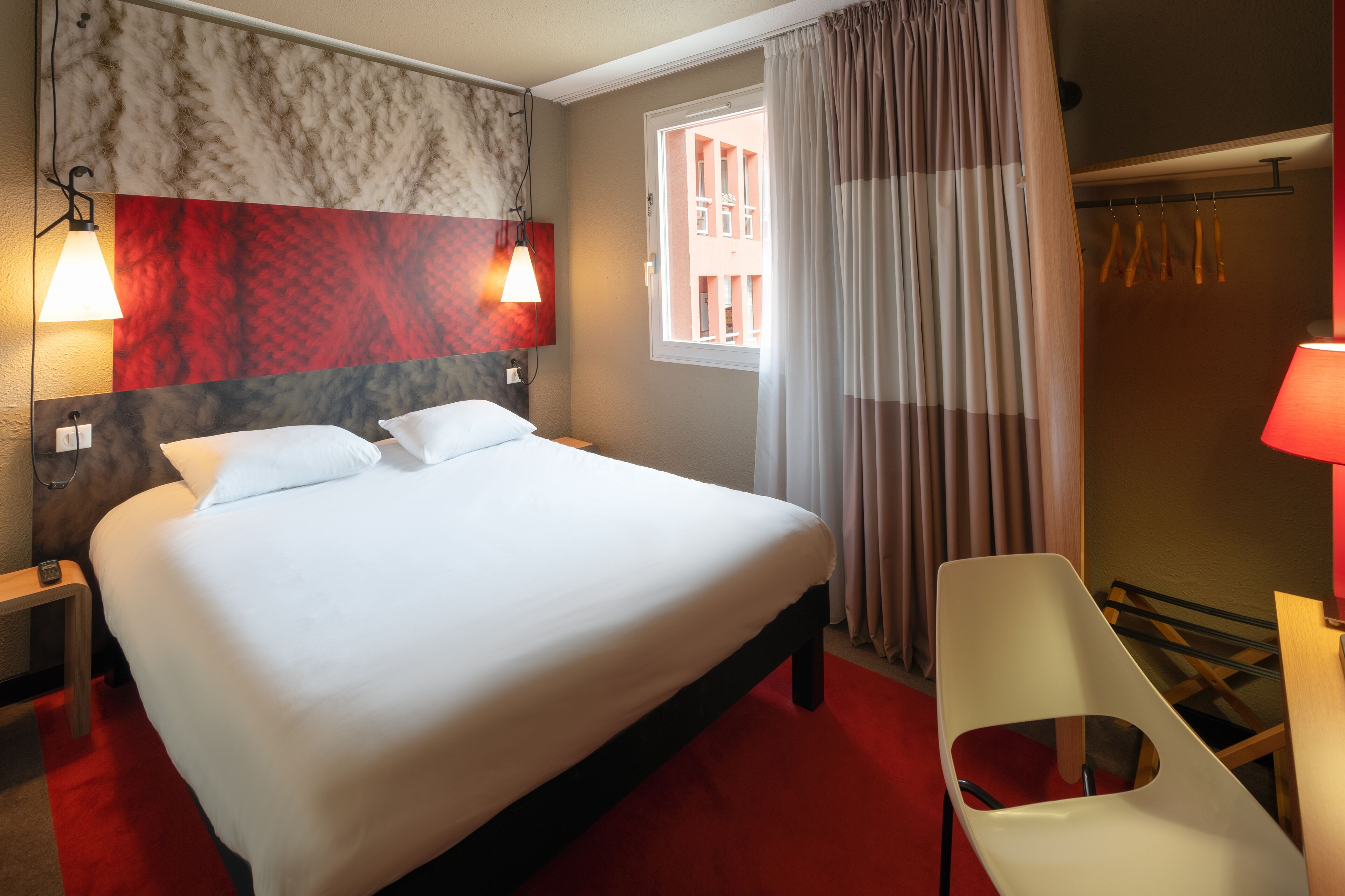 Una habitación de hotel con cama, lámparas de noche, ventana con cortinas, una silla y zona de ducha.