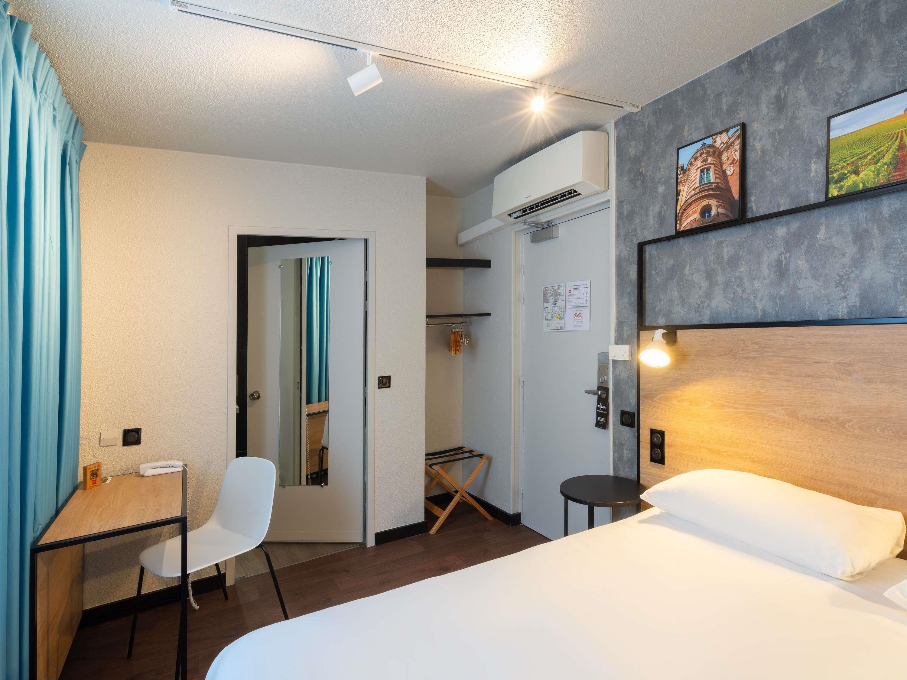 A hotel room with a bed, a desk, a chair, an air conditioning unit, and a door with a mirror.