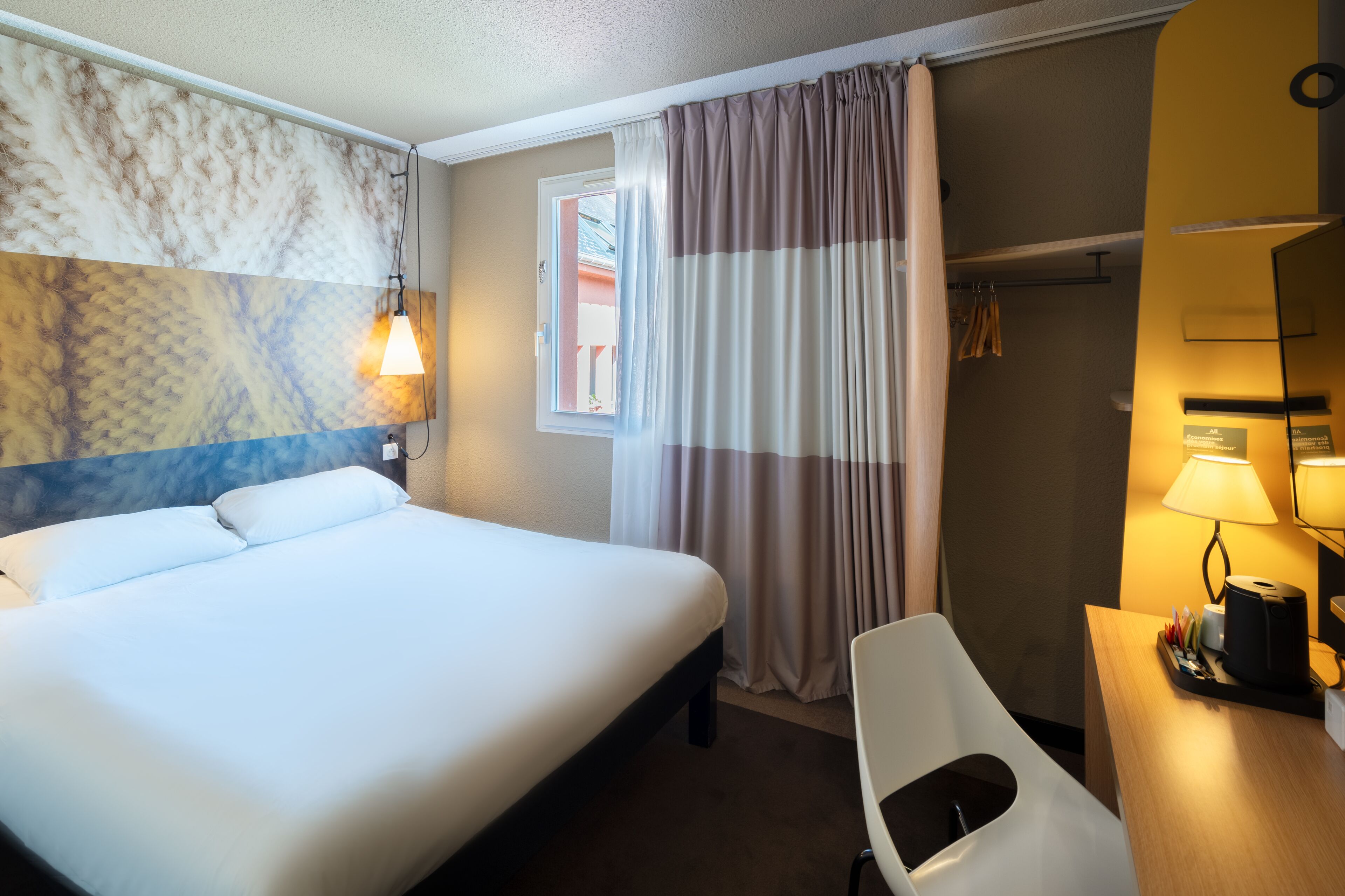 Habitación de hotel con cama, escritorio con lámpara, silla, ventana con cortinas y pared con diseño texturizado.