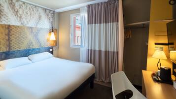 Habitación de hotel con cama, escritorio con lámpara, silla, ventana con cortinas y pared con diseño texturizado.