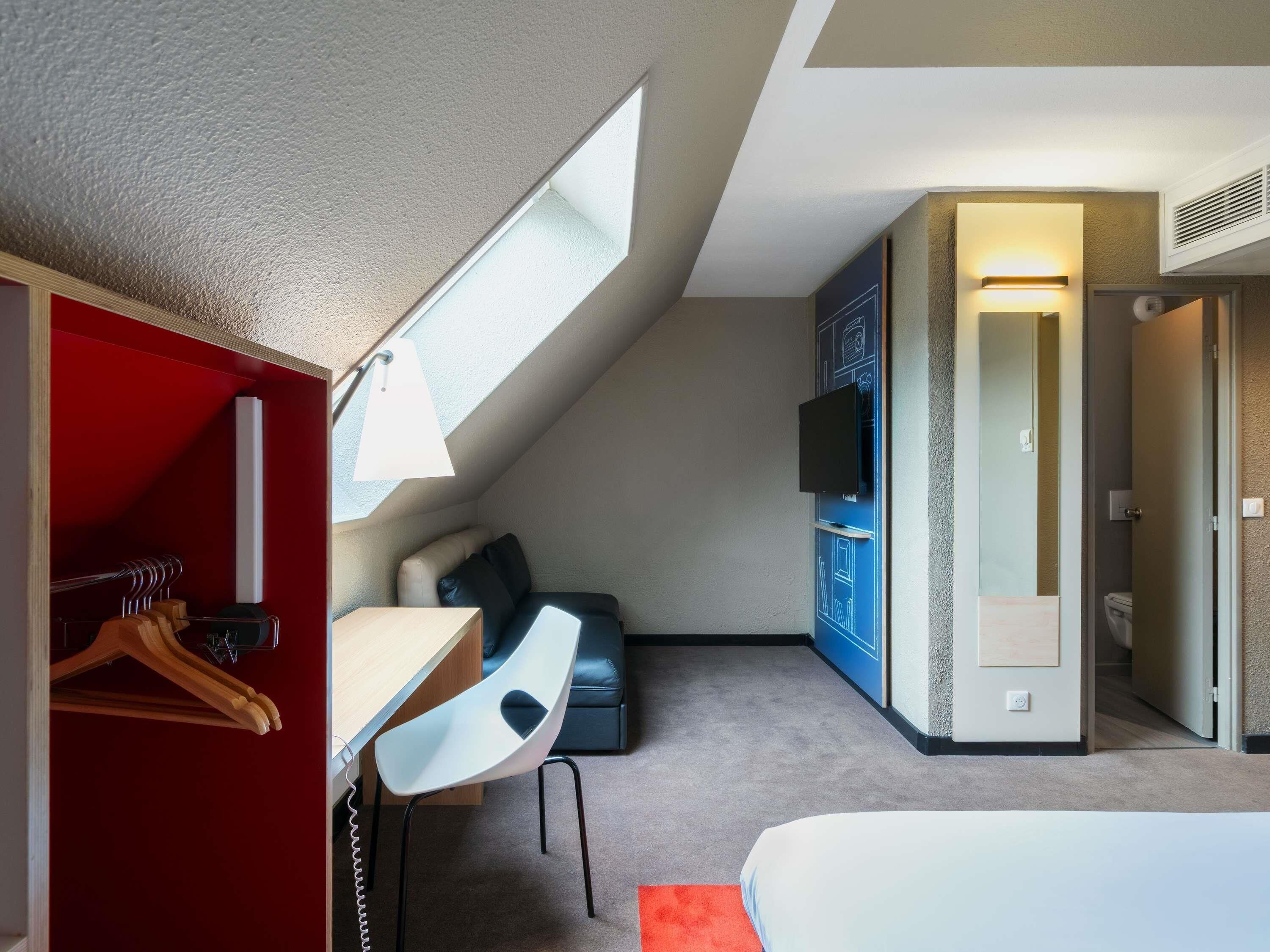 Habitación de hotel con cama, escritorio con silla, televisor montado en la pared y un pequeño rincón de estar con sofá y almohadones.
