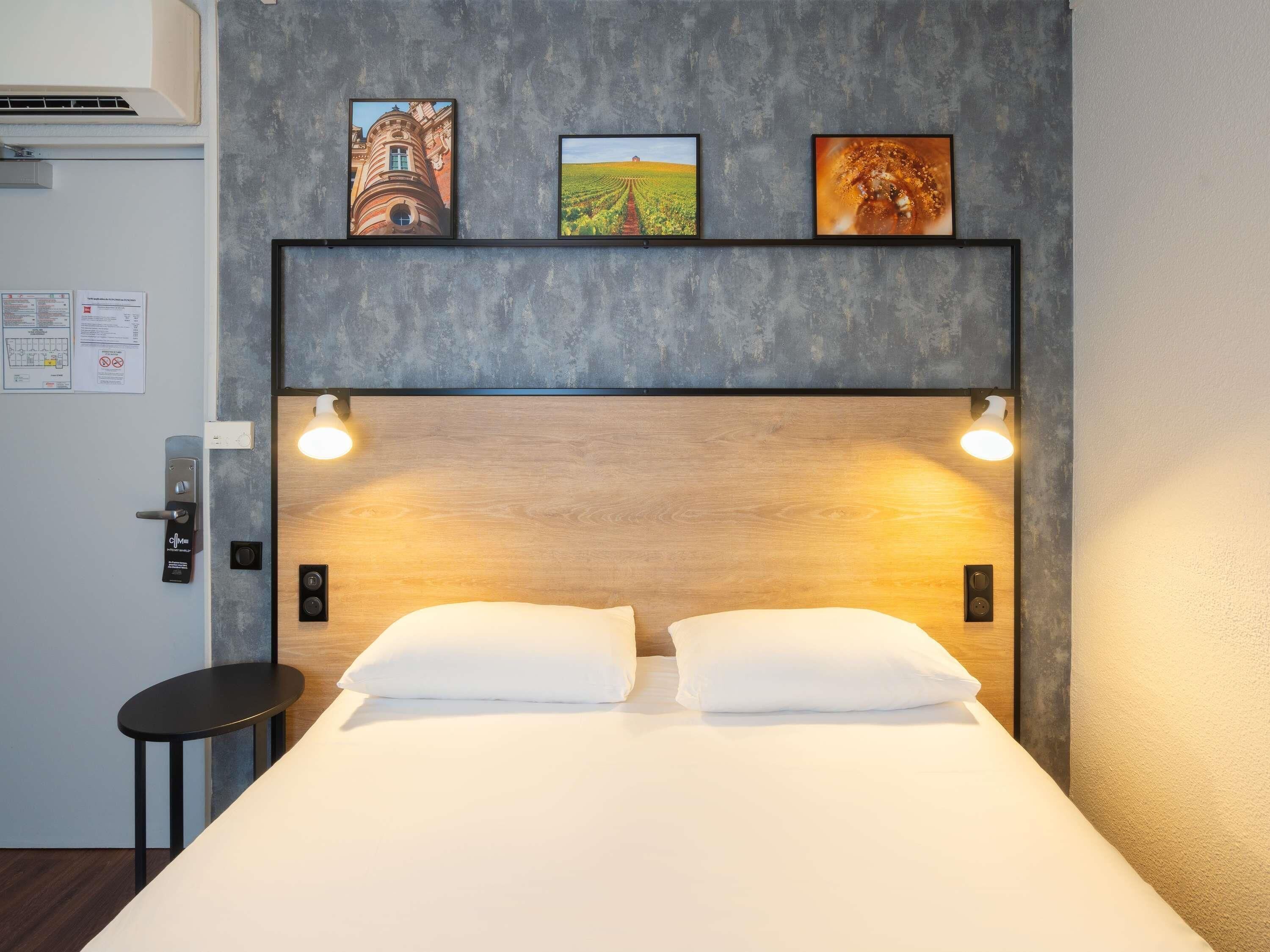 A hotel room with a bed, two bedside lamps, a wooden headboard, and three framed pictures on the wall.