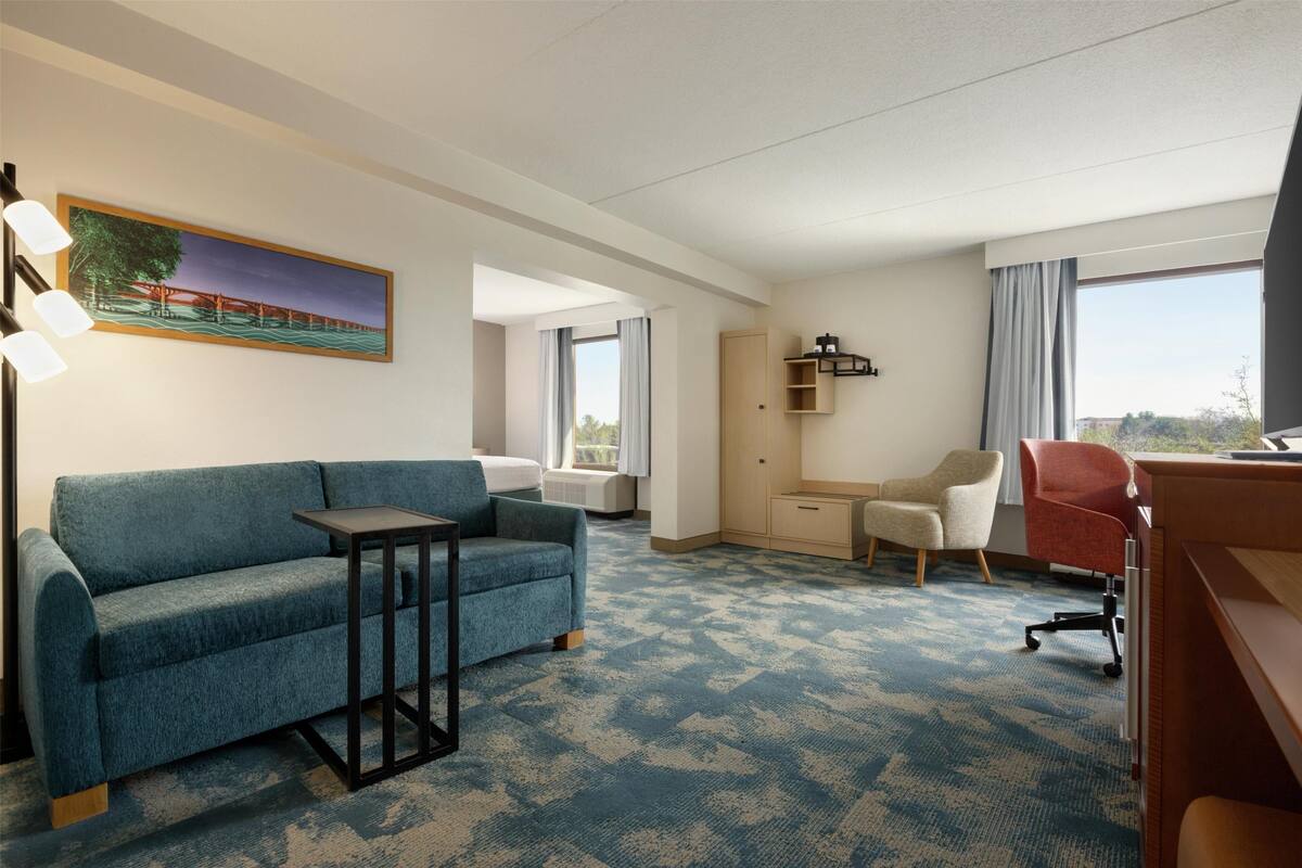 A hotel room with a blue sofa, a small table, a desk with a chair, a wardrobe, and a painting on the wall.