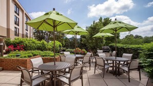 Eine Terrasse im Freien mit runden Tischen und Stühlen, grünen Sonnenschirmen und einem Ziergarten aus Ziegelsteinen mit Blumen.