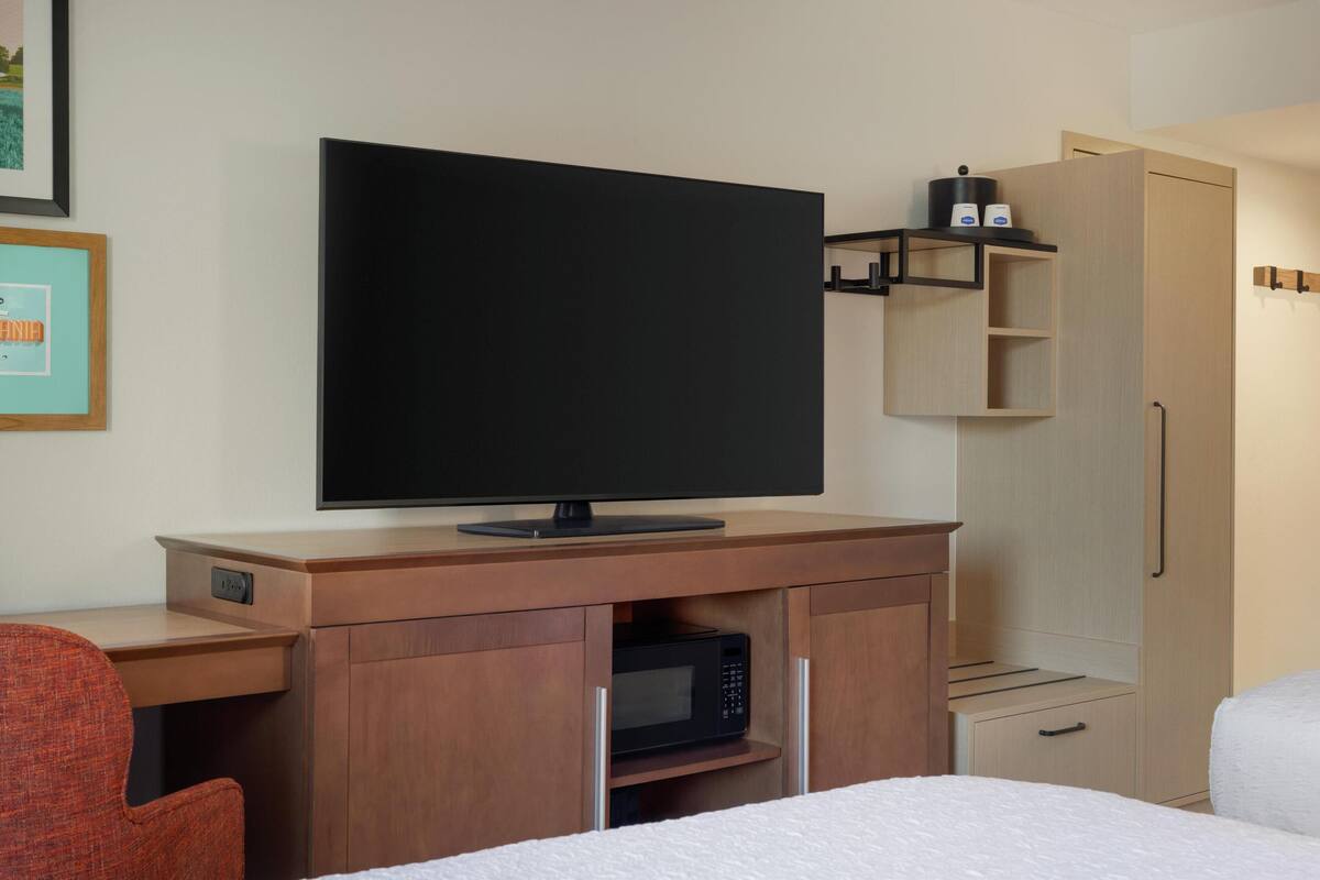 A hotel room with a flat-screen TV on a wooden desk, a microwave, a wardrobe, and a bed with white bedding.