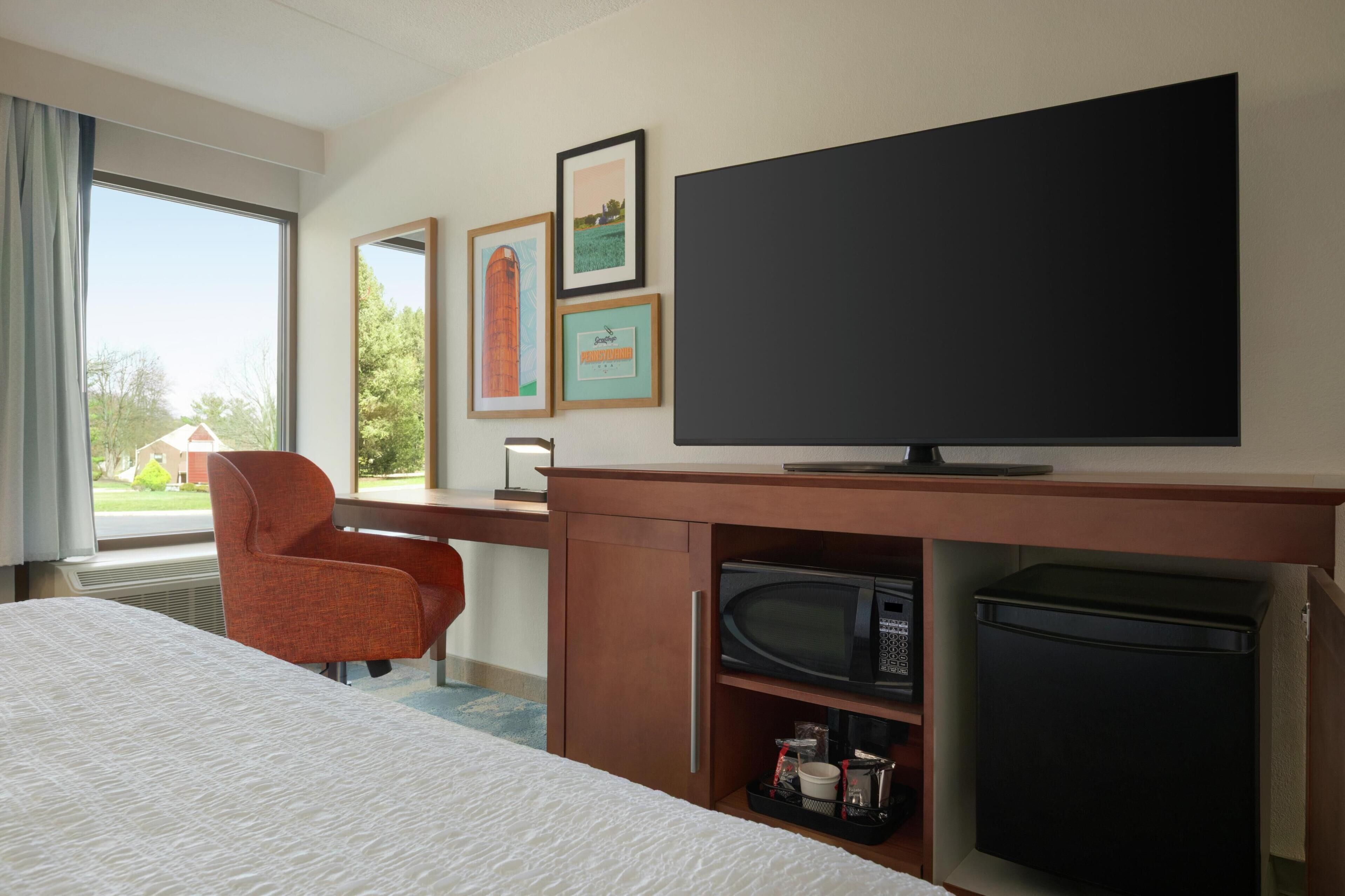Quarto de hotel com cama, escrivaninha com TV de tela plana, micro-ondas, geladeira e uma cadeira. Há quadros nas paredes.