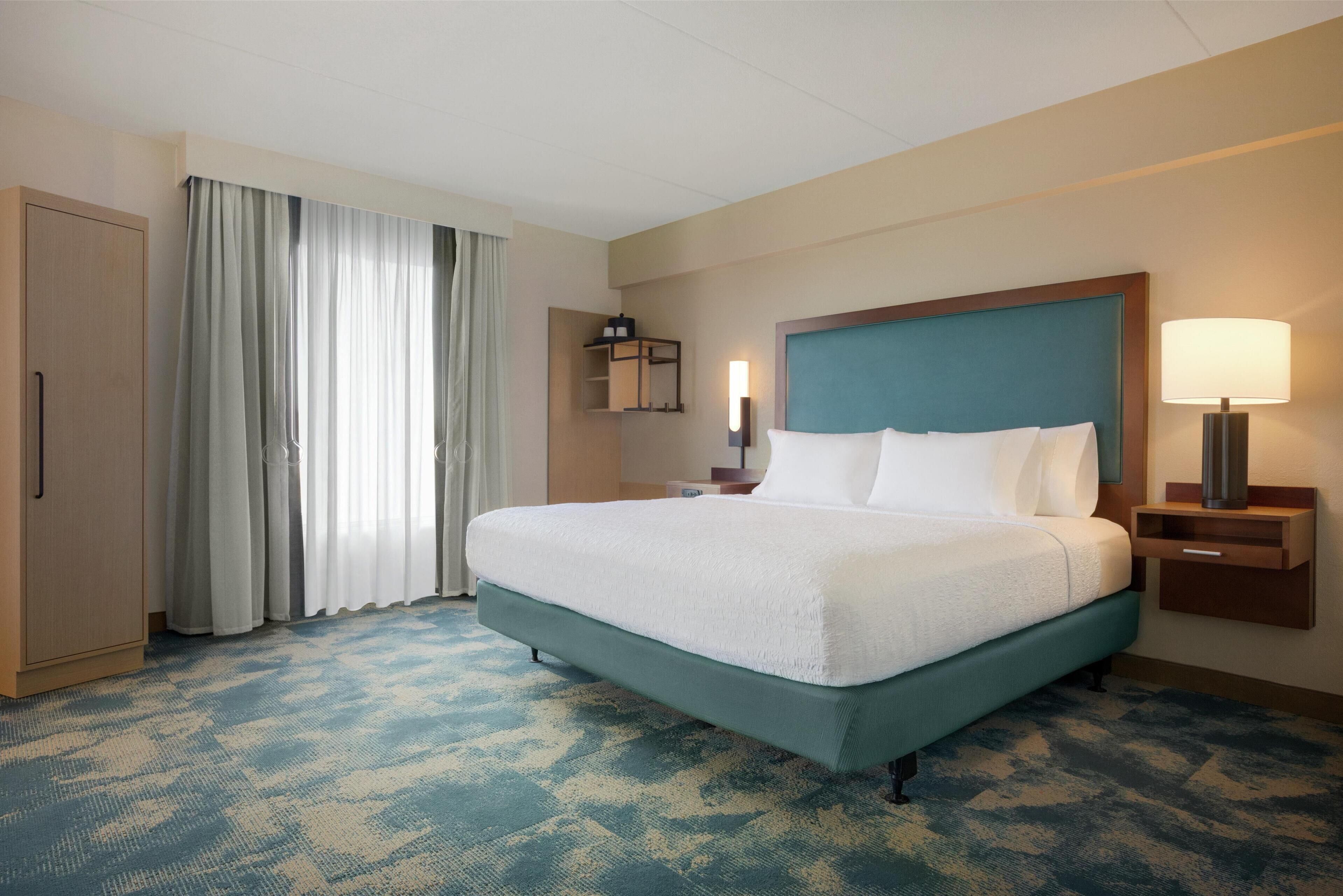 A hotel room with a large bed, a nightstand, a lamp, a wardrobe, and a window with curtains.