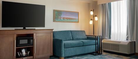 A hotel room with a large flat-screen TV, a sofa, a cabinet with a microwave, a painting on the wall, and a window with curtains.