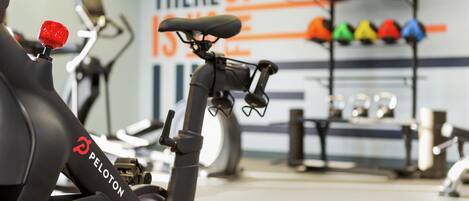 Ein stehendes Fahrrad mit einem roten Knopf und dem Peloton-Logo, in einem Fitnessstudio mit einer motivierenden Wand und bunten Kurzhanteln.