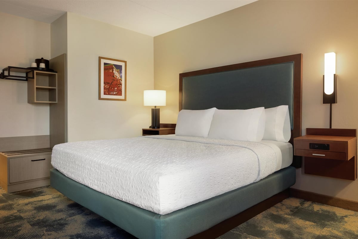 A hotel room with a large bed, a bedside table with a lamp, a small shelf, and a framed picture on the wall.