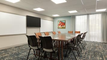 Un salón de conferencias con una larga mesa, sillas, una pantalla de proyector, un cuadro y una ventana con cortinas.