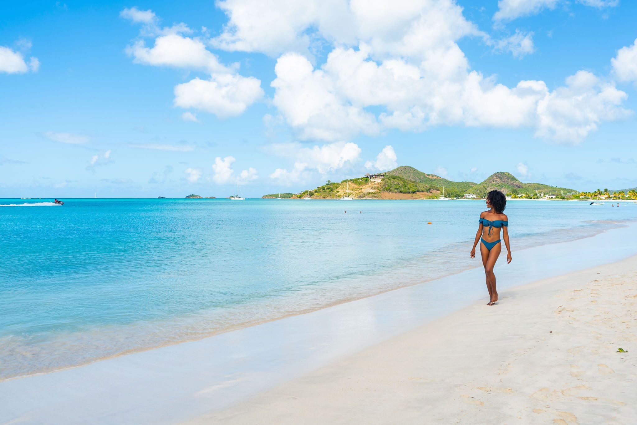 Una mujer en traje de baño caminando por una playa arenosa con agua azul cristalina y colinas al fondo.