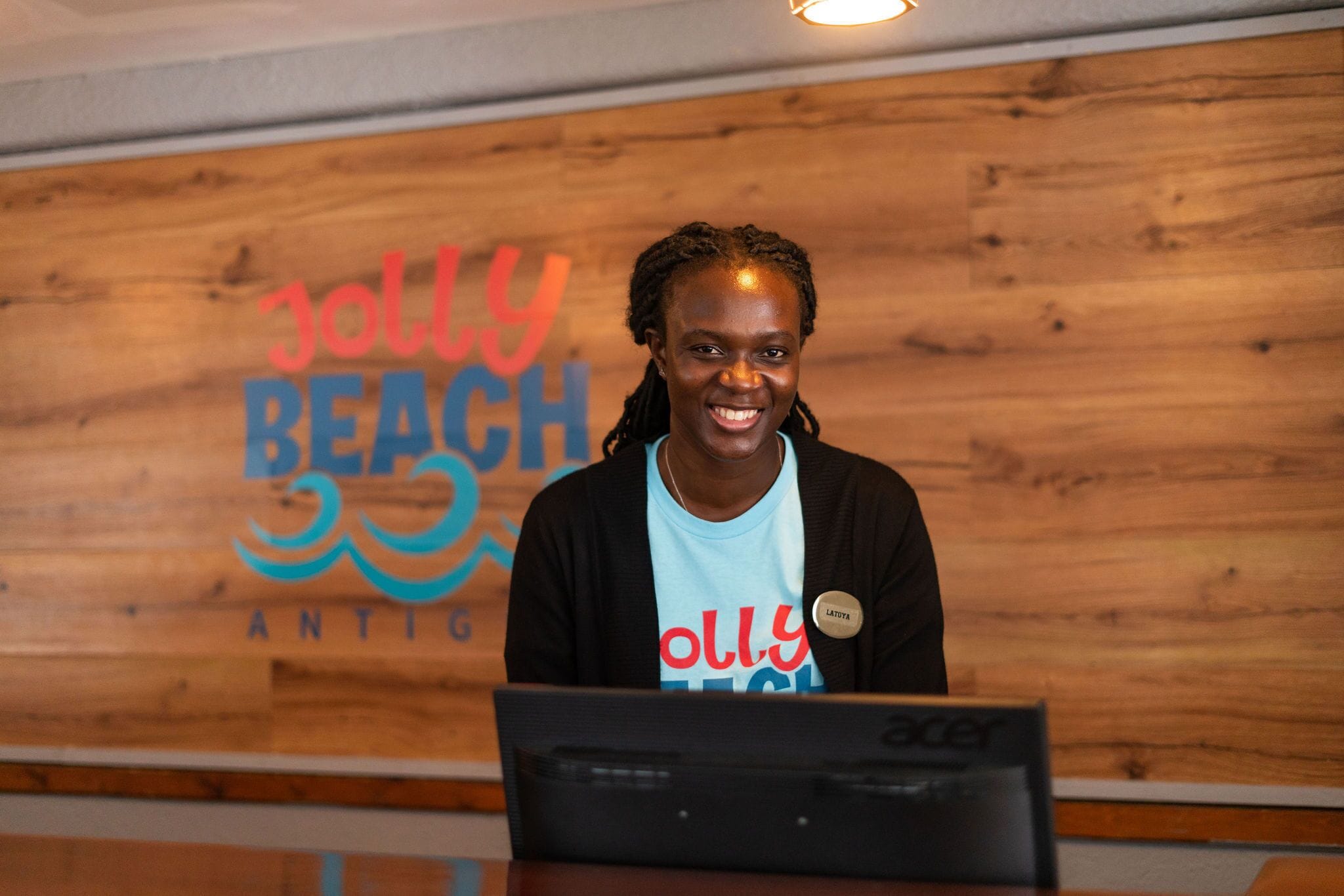 Una persona sentada en un escritorio con una computadora portátil frente a una pared de madera con el texto 'Jolly Beach Antig'.
