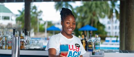 A person at a bar with a drink, a bar counter, and a view of palm trees and a building in the background.