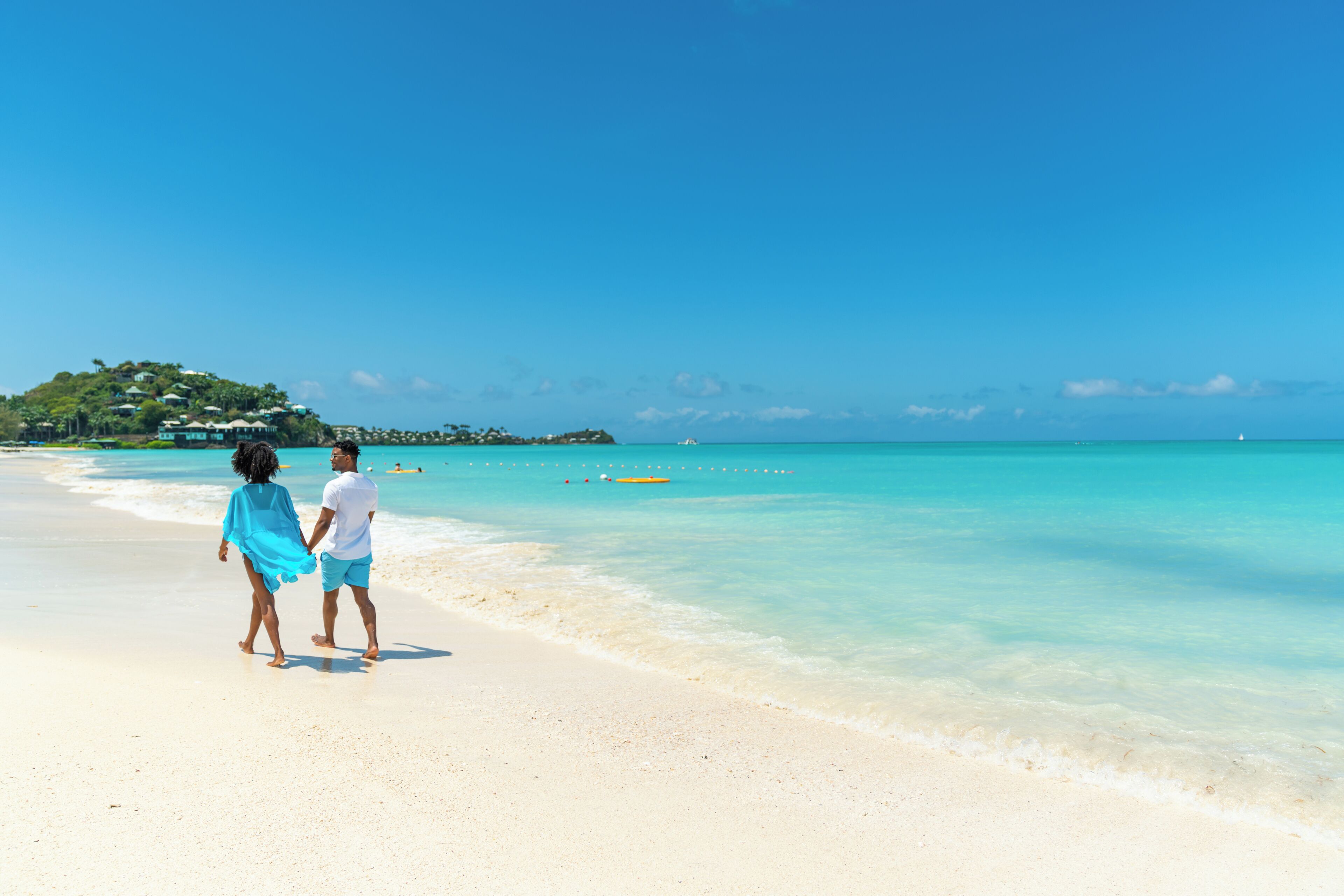 Una playa con agua turquesa cristalina, una pareja caminando sobre la arena, y una vista distante de una pequeña isla con edificios.
