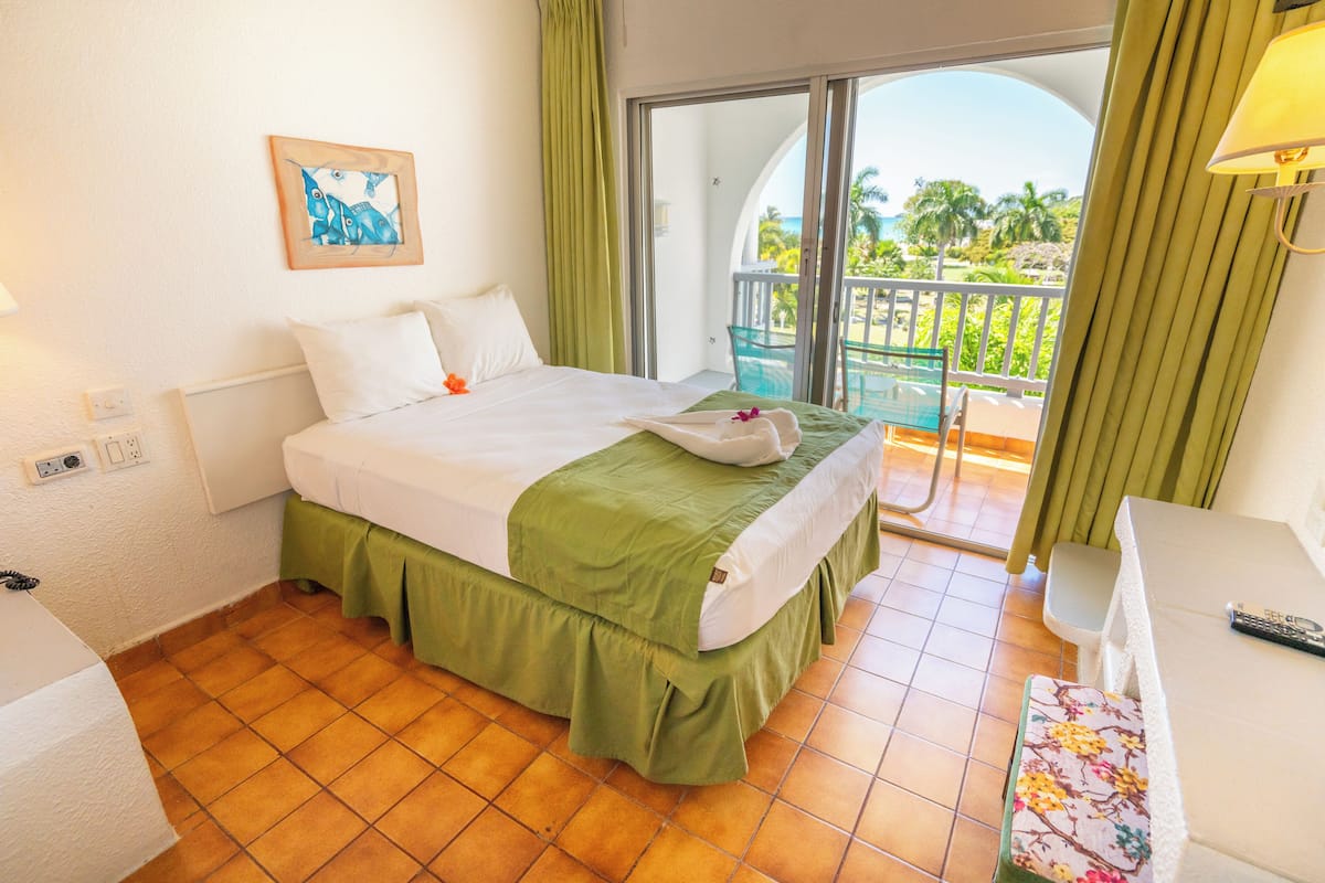 A hotel room with a bed, a balcony with a view, a painting on the wall, and a desk with a lamp.