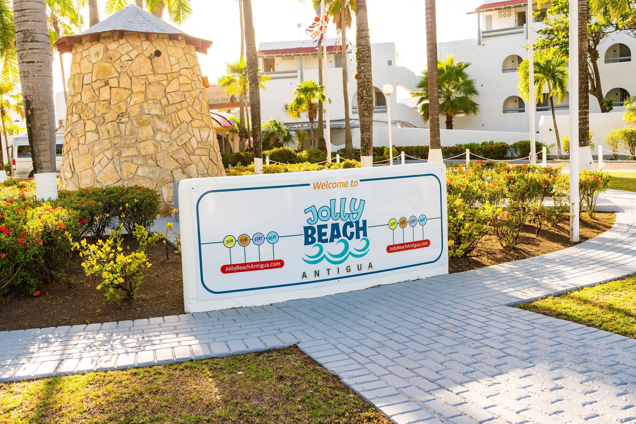 Welkomstbord voor Jolly Beach in Antigua, met een stenen structuur, palmbomen en een verharde pad.