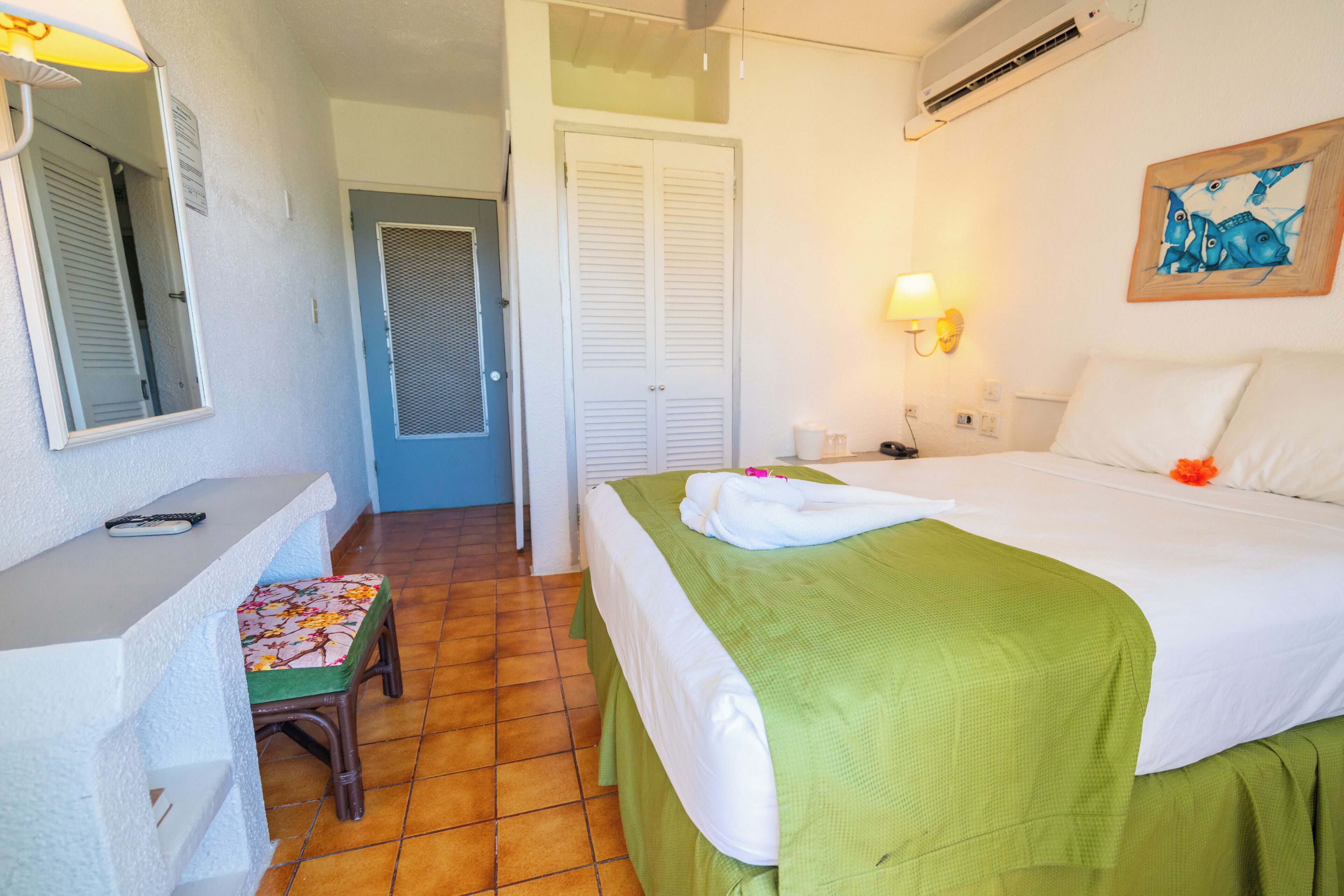 A hotel room with a bed, a desk, a chair, a mirror, a door, and a wall-mounted air conditioner.