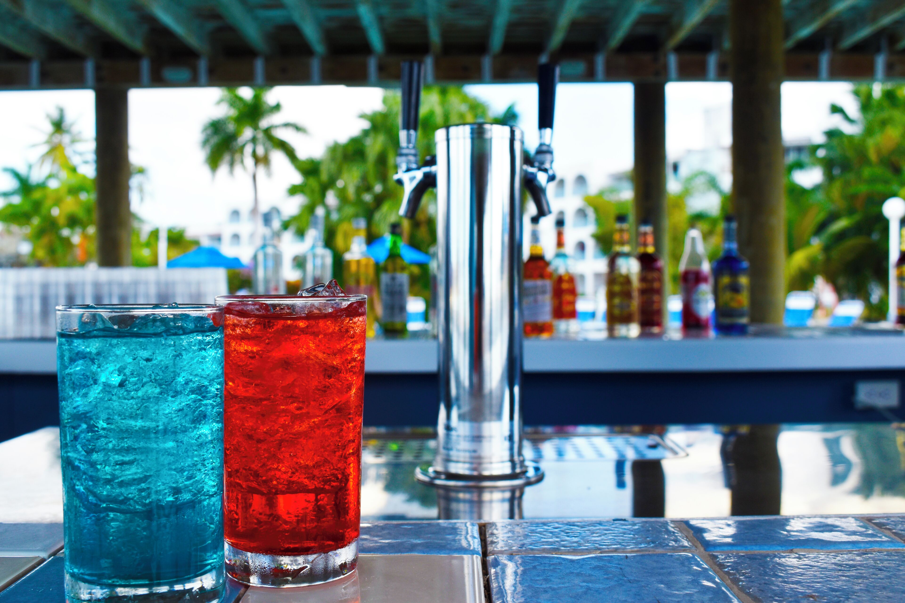 A bar area with two glasses of iced drinks, a metal tap, and a view of palm trees and a pool in the background.