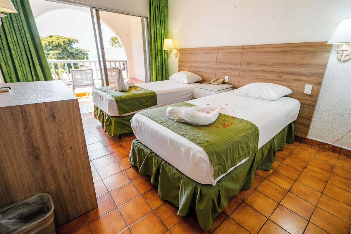 A hotel room with two beds, a wooden desk, a trash bin, and a balcony with a view.