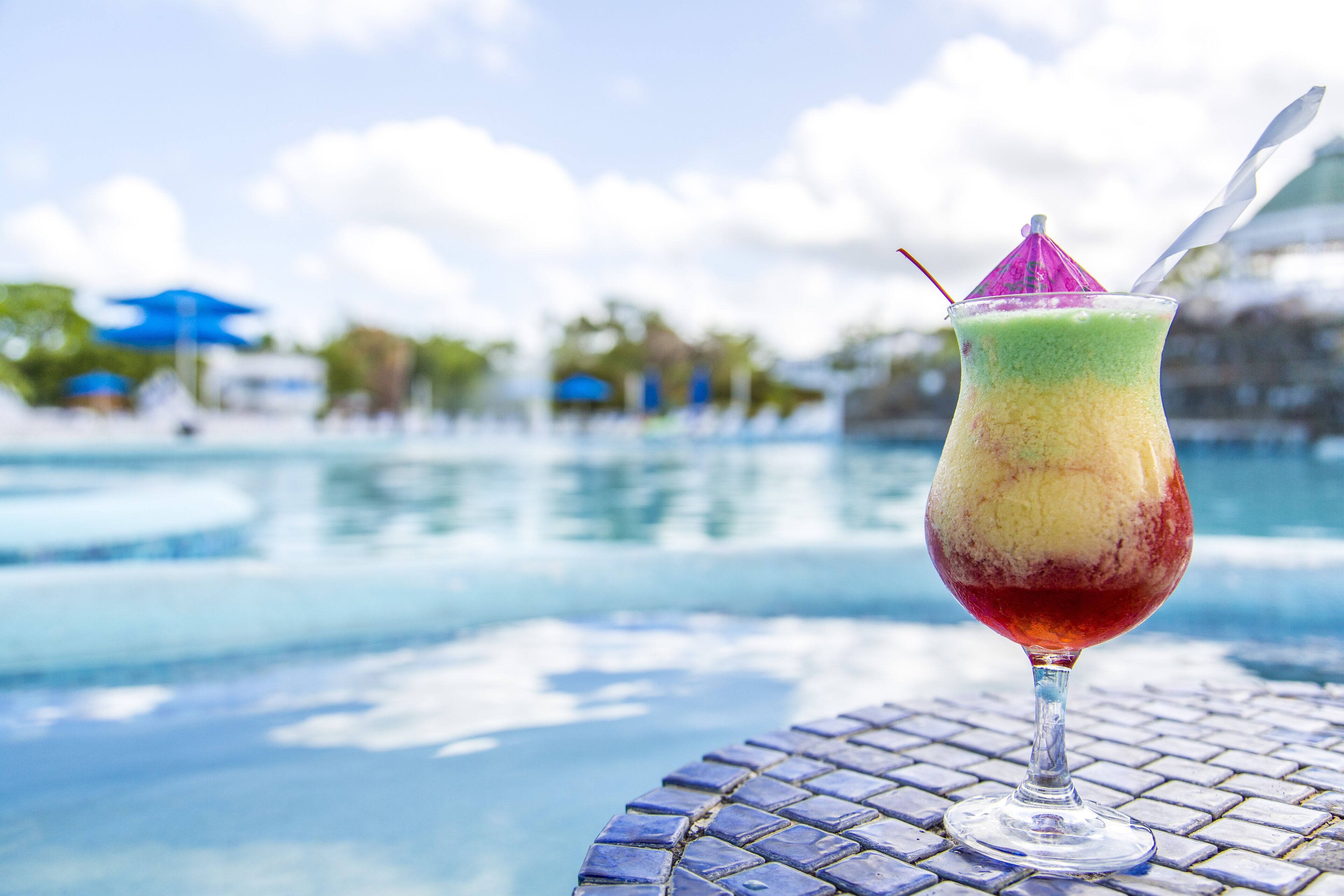 A colorful drink in a glass with a decorative umbrella on a table by a pool.