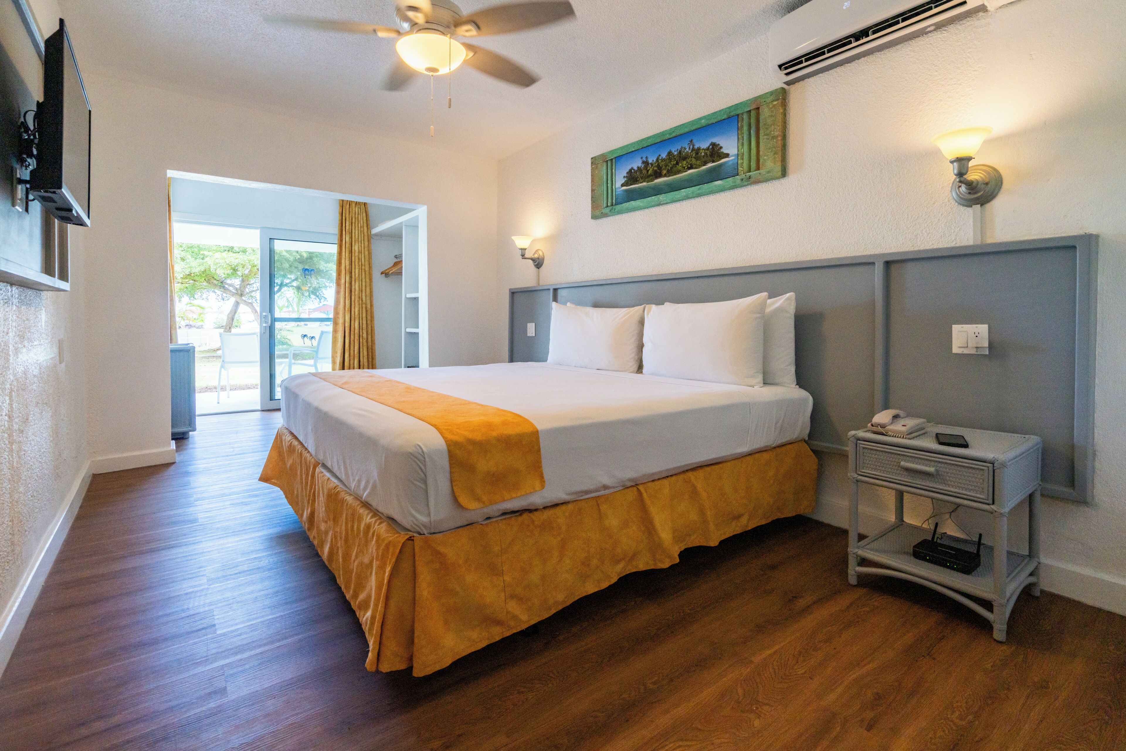 Une chambre à coucher avec un lit, une table de chevet, une commode, un ventilateur de plafond et un tableau accroché au mur.