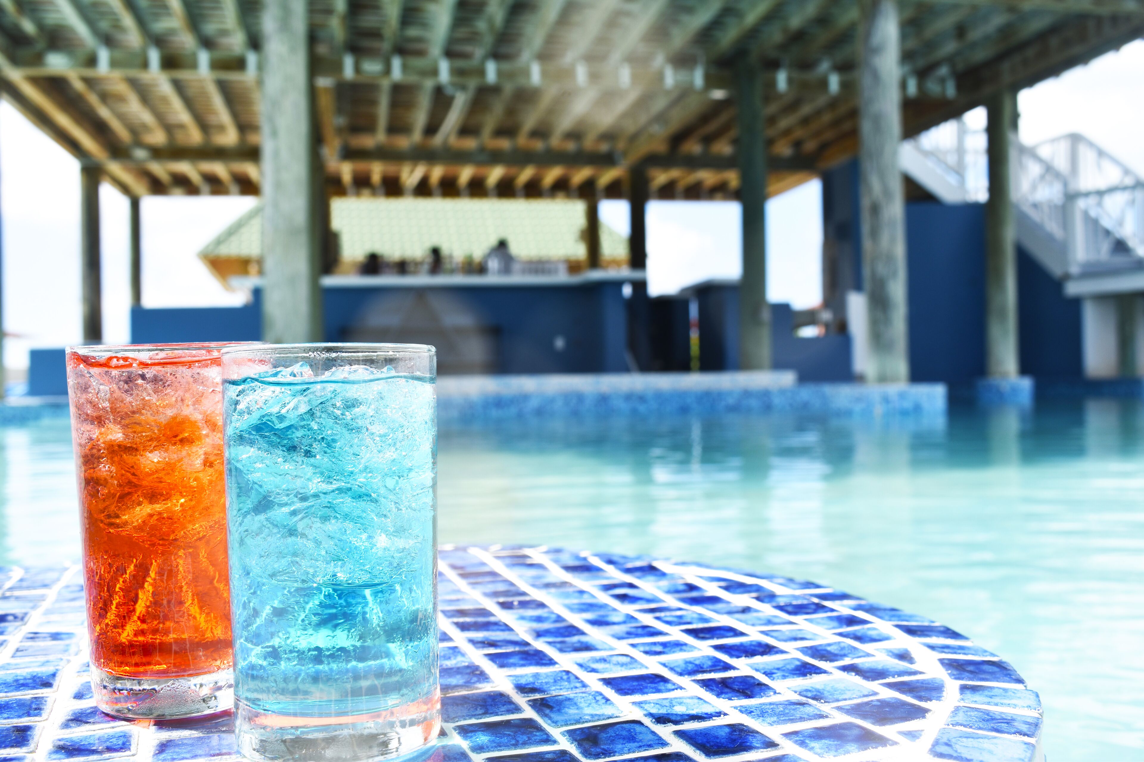 Two glasses of iced beverages on a tiled table by a poolside.