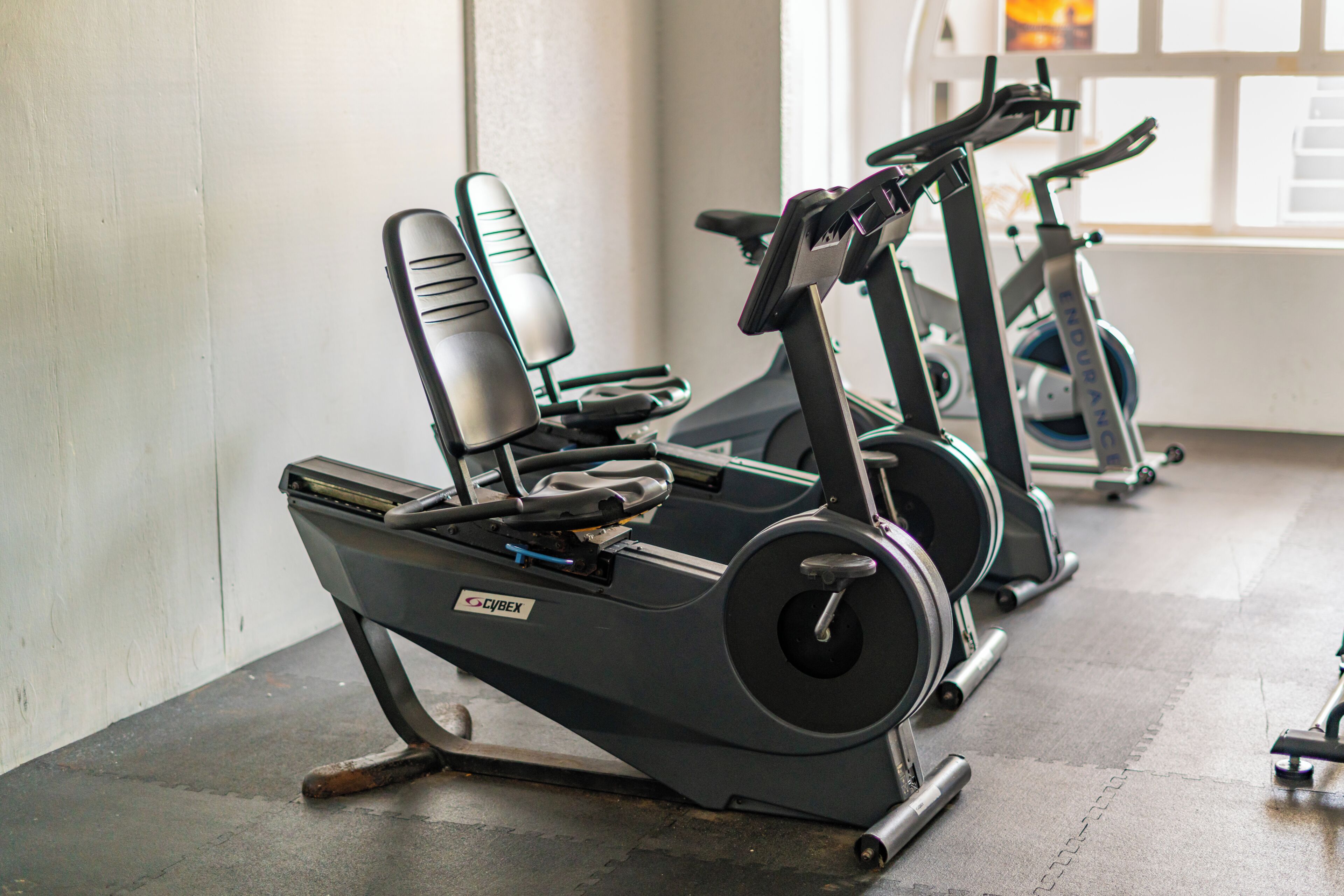 Een sportschool met hometrainers en loopbanden, een betonnen vloer en een raam met daglicht.