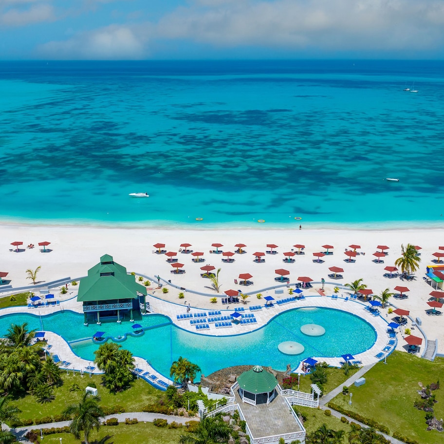 An aerial view of a resort with a large swimming pool, beach umbrellas, and a clear view of the ocean.