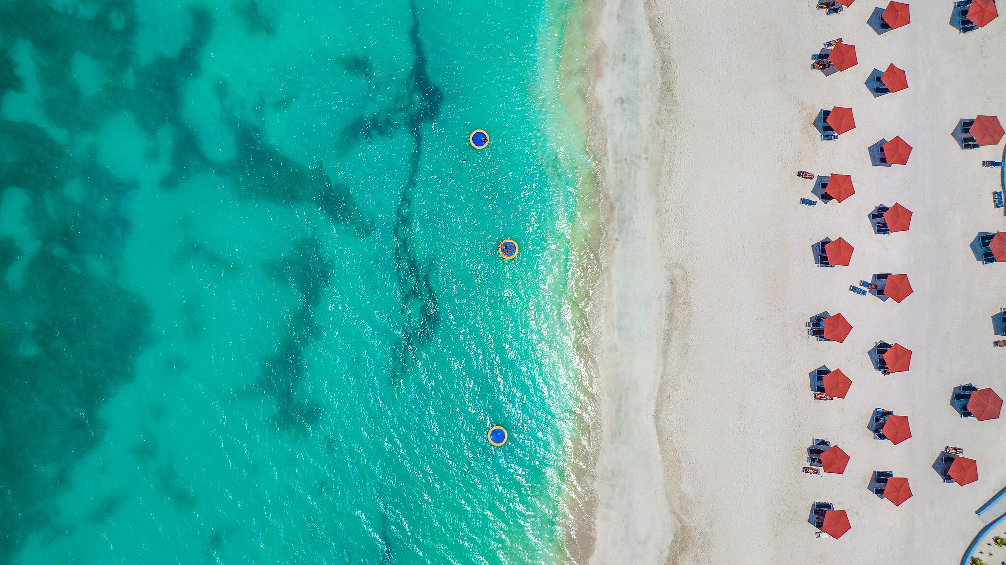 Ovenudblik over en strand med klart turkis vand, en sandstrand og flere røde parasoller arrangeret i rækker.