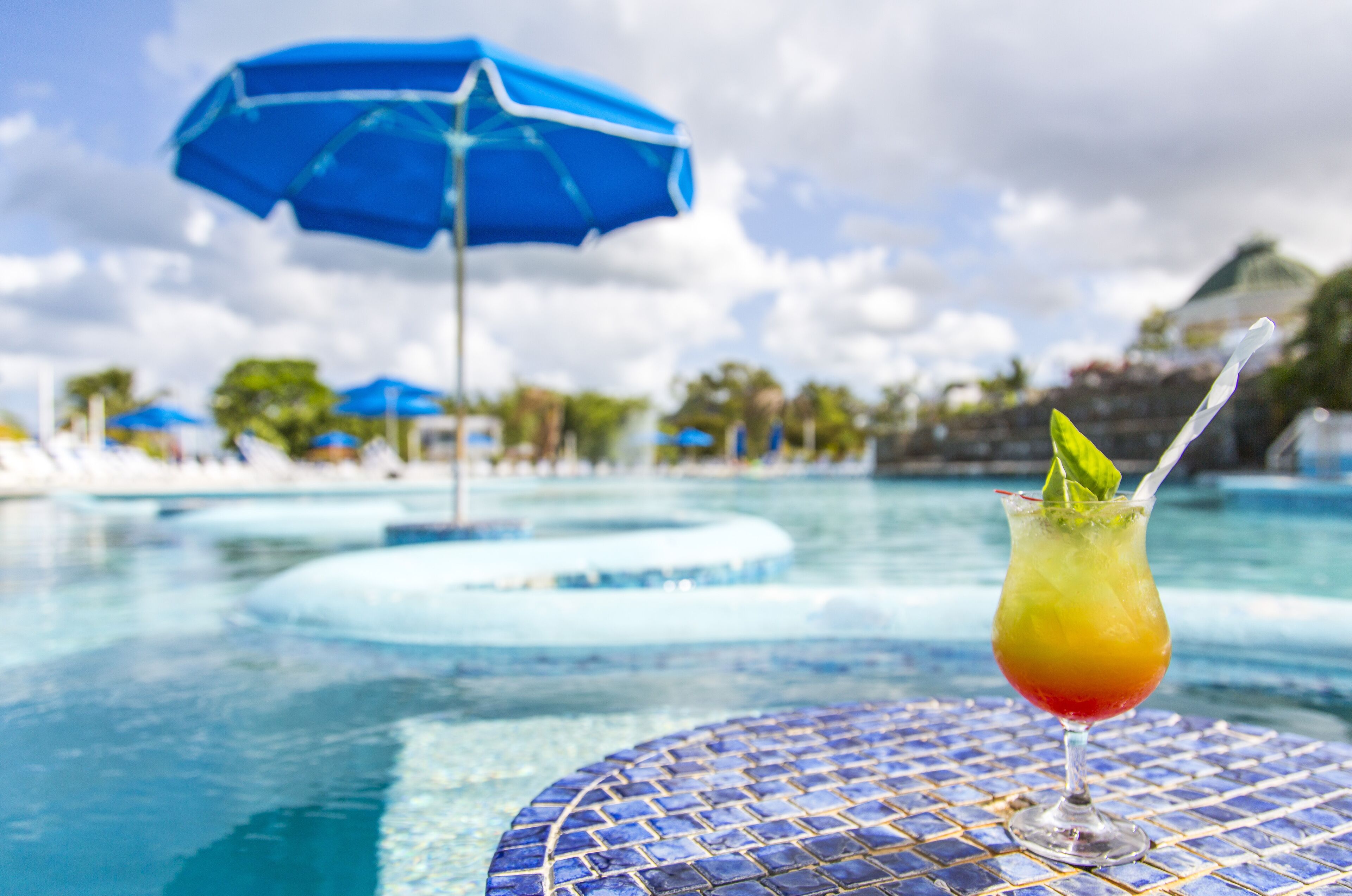 A poolside view with a drink in a glass, a blue umbrella, and a tiled table.