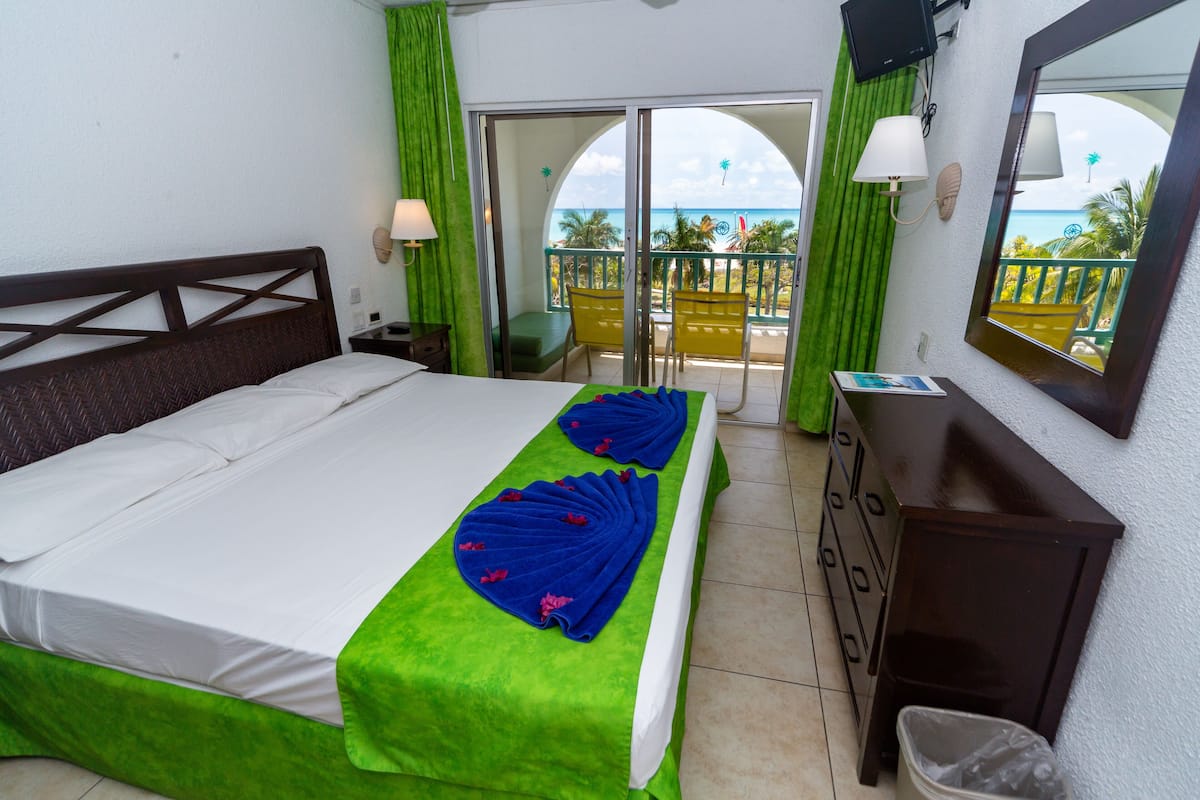 A hotel room with a bed, a dresser, a mirror, a balcony with a view of the ocean, and a door leading to the balcony.