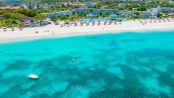Vista aérea de un complejo turístico en la playa, con un hotel grande, aguas turquesas cristalinas y una playa de arena con sombrillas.
